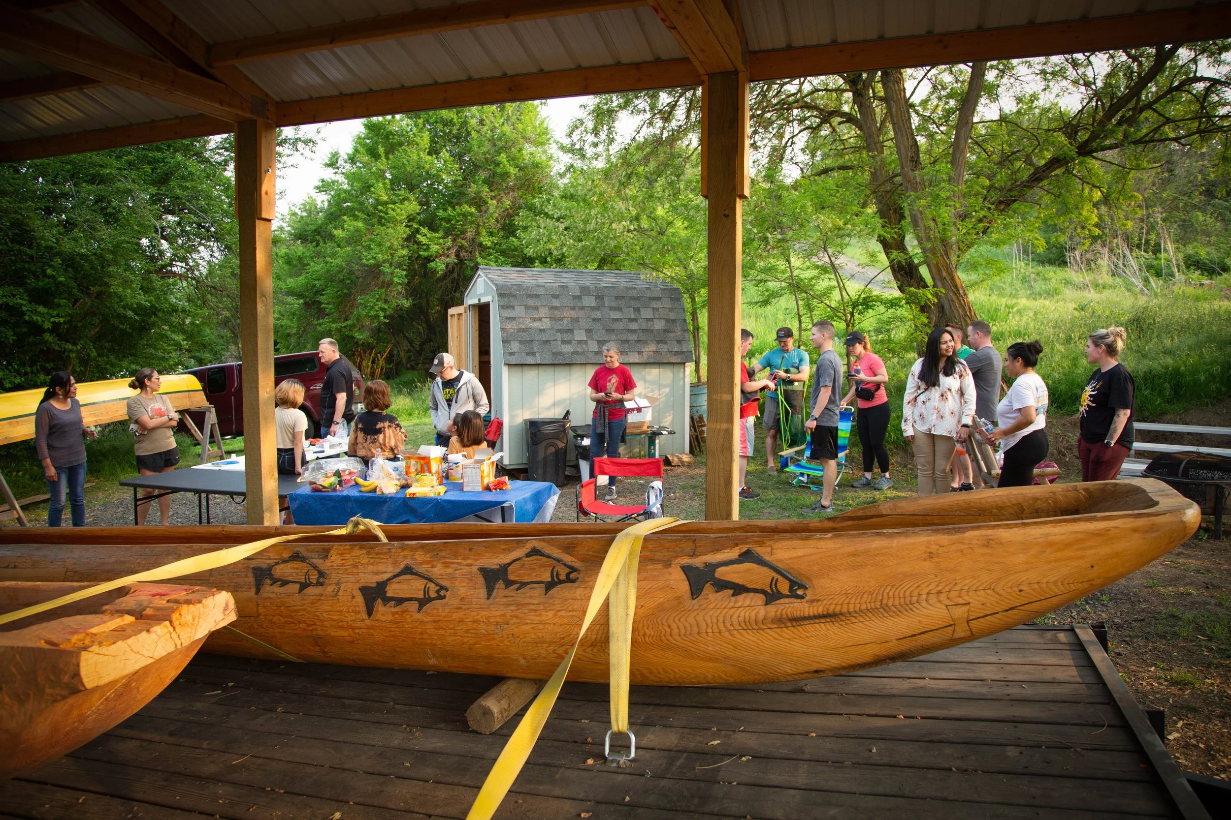 People gathered outdoors during a gathering or event, seen through a large wooden canoe resting on a wooden platform, with trees and greenery in the background.