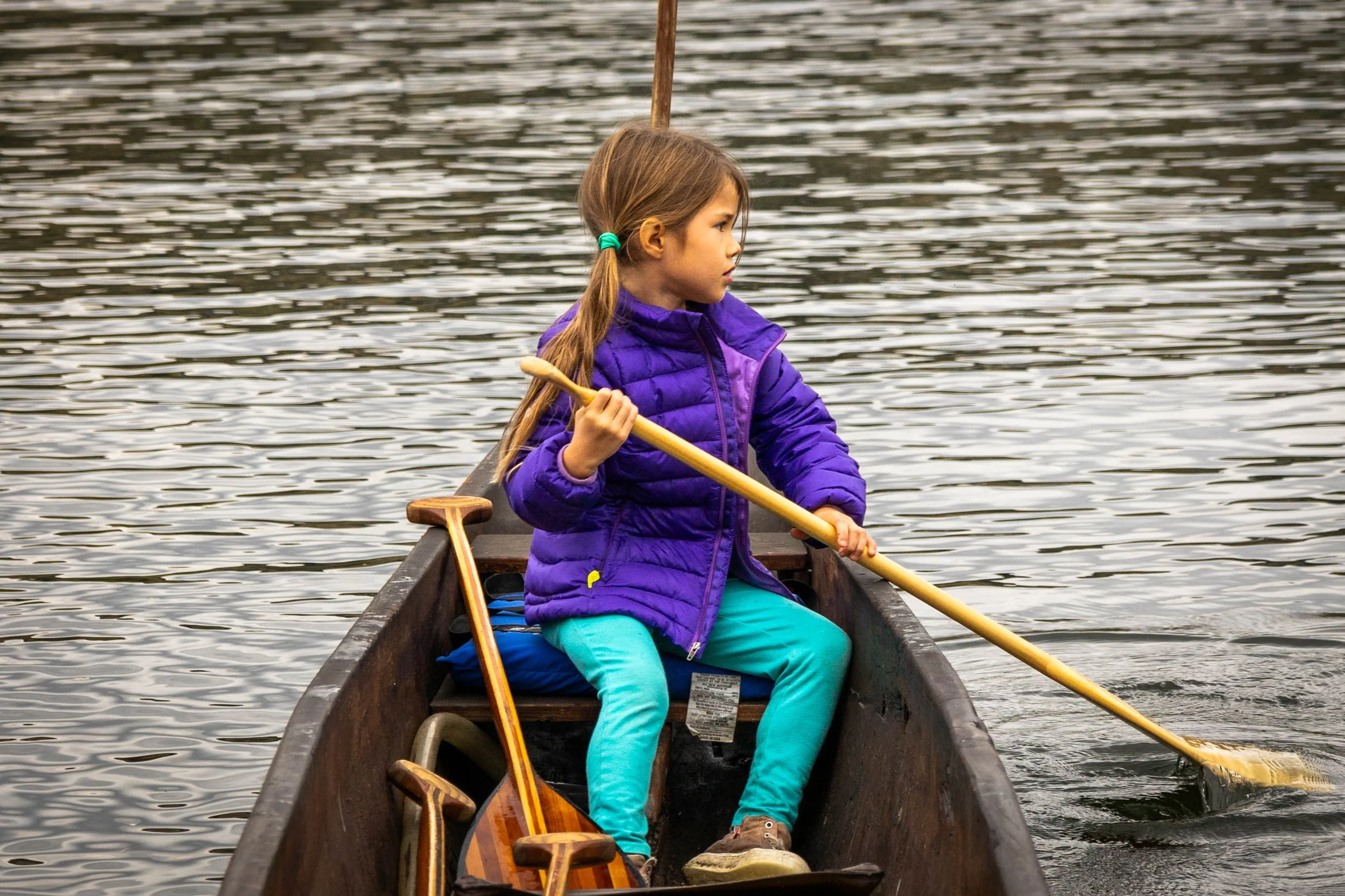 A young girl with long hair tied in a ponytail, wearing a purple jacket and blue-green pants, sitting in a canoe on a body of water. She is holding a paddle and looking to the side.