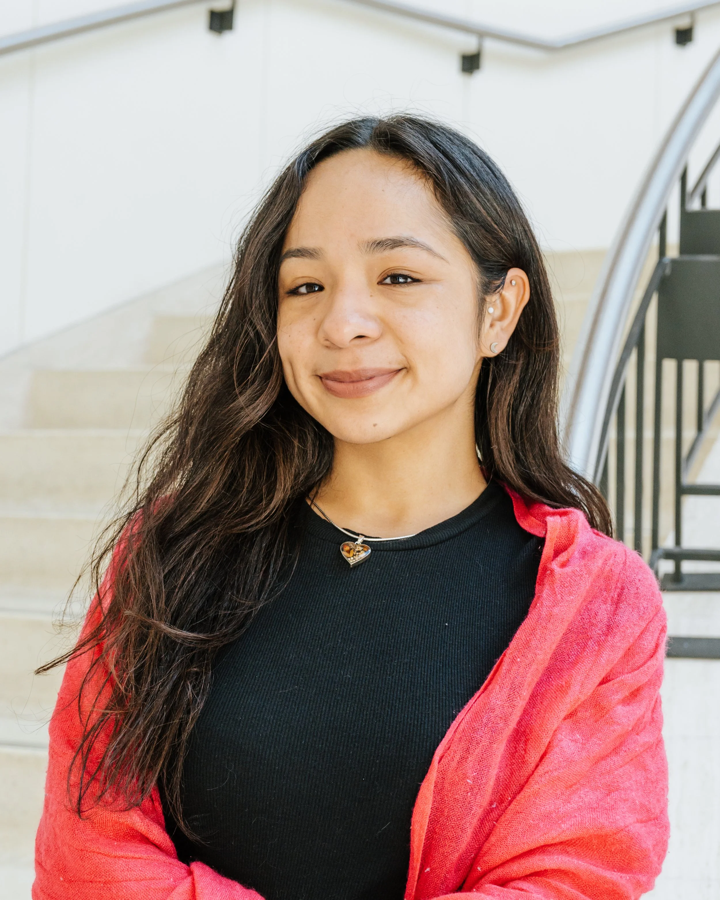 Close-up of a young woman with long, dark hair, smiling softly, wearing a black top and red jacket, with stairs in the background.