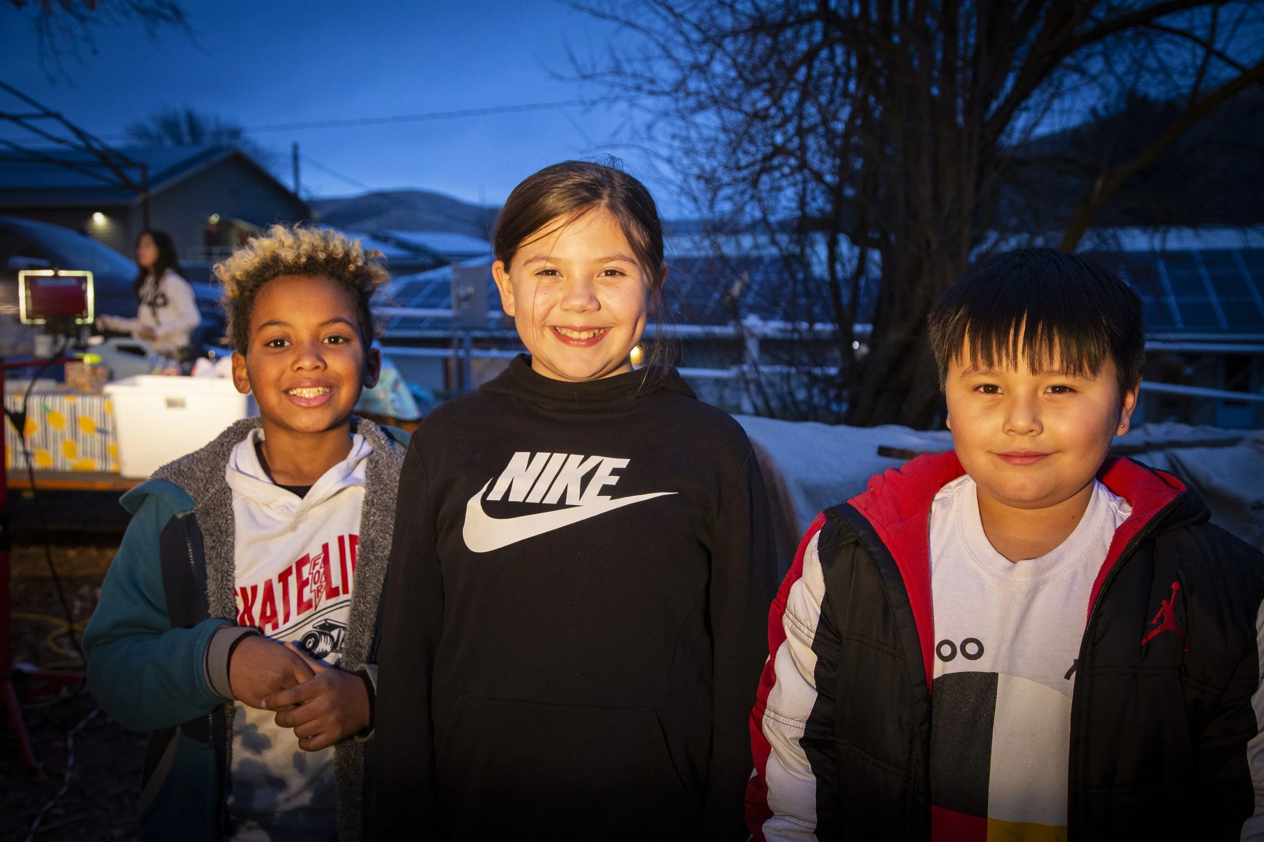 Three children standing outdoors during dusk, smiling at the camera, with a girl in a white and red Nike sweatshirt on the left, a girl in a black Nike sweatshirt in the middle, and a boy in a red and white jacket on the right.
