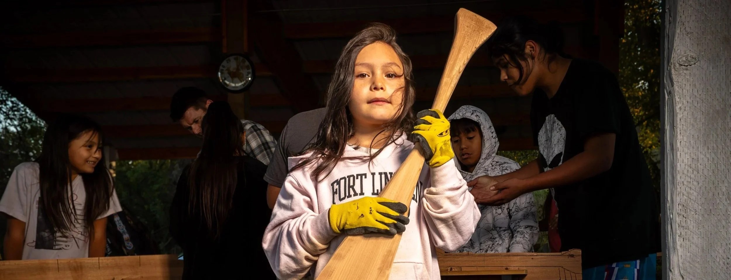 Young girl with long brown hair holding a wooden bat and wearing yellow gloves, standing among other children and adults during an outdoor campfire or group activity at dusk.