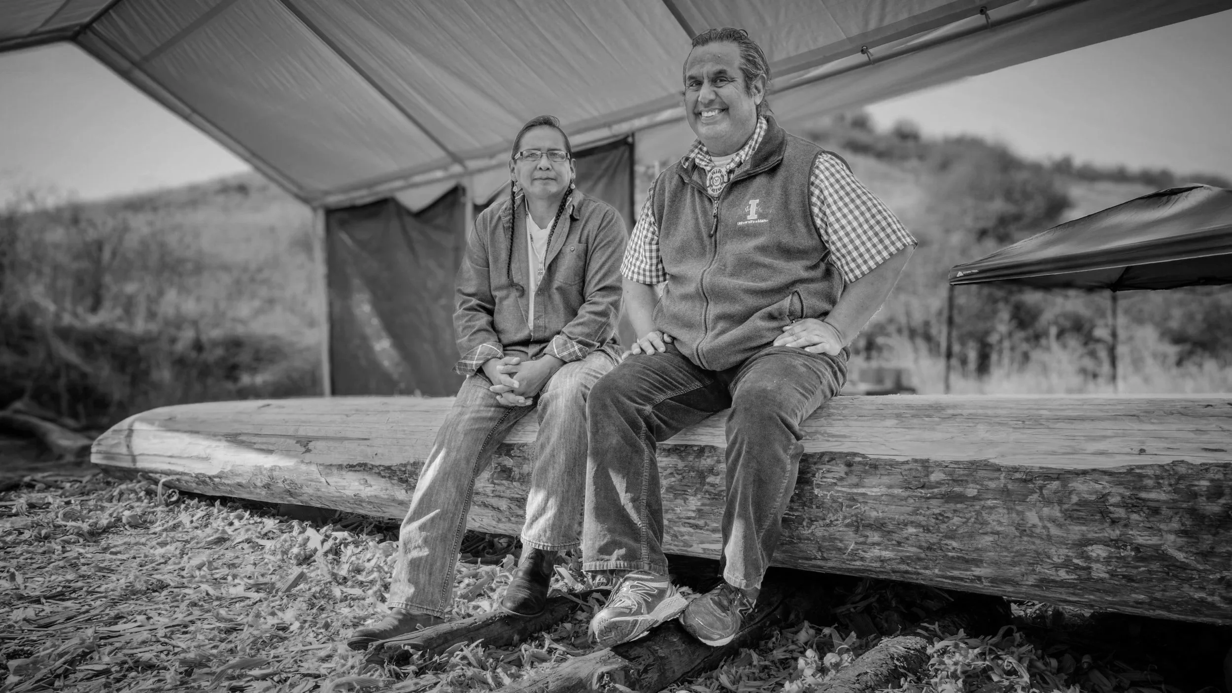 Two people sitting on a large piece of wood outdoors under a canopy, with trees and hills in the background. They are smiling and looking at the camera.