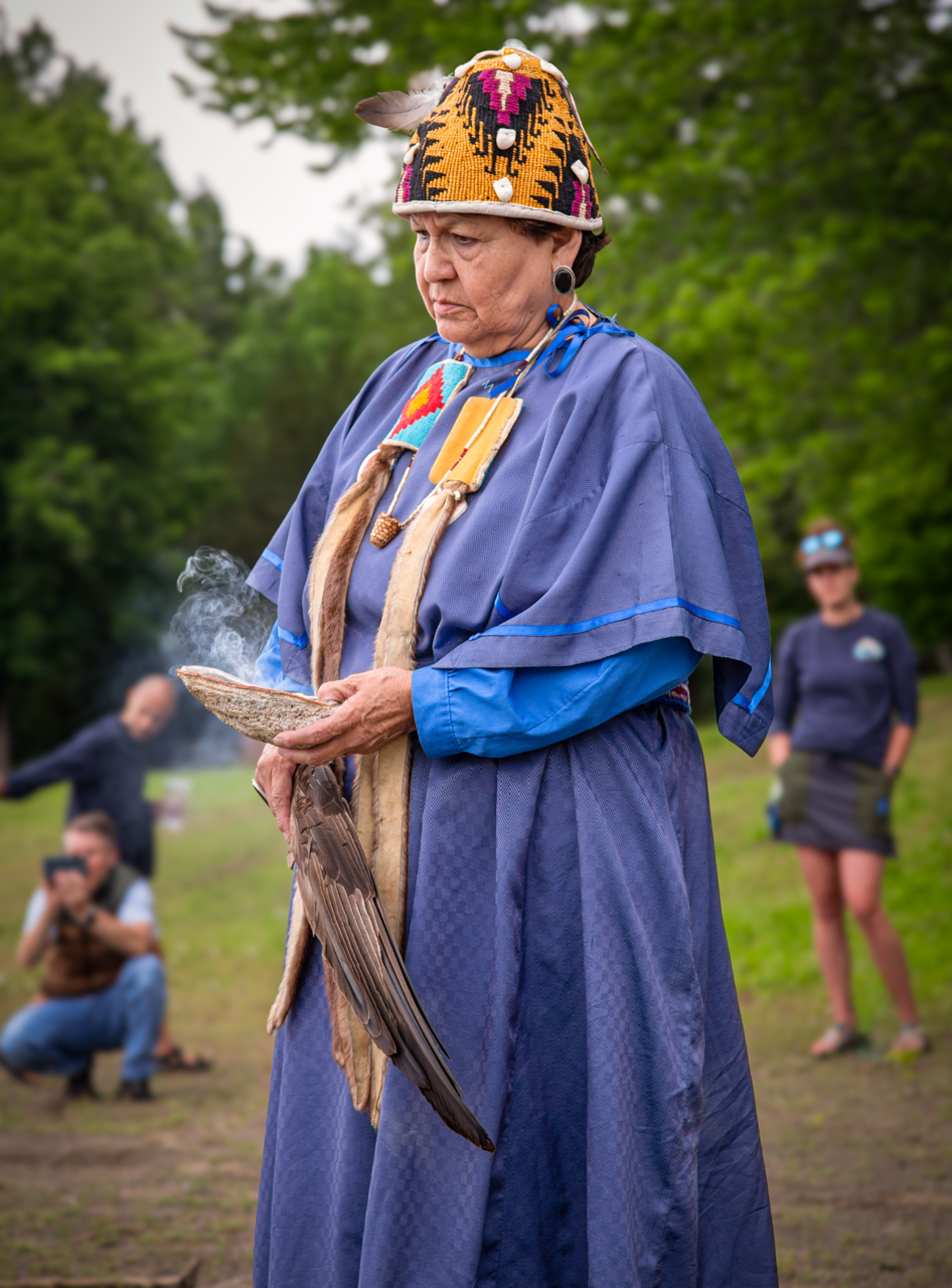 Elder woman in traditional indigenous attire, holding a smoking ceremonial object, outdoors with trees in the background.