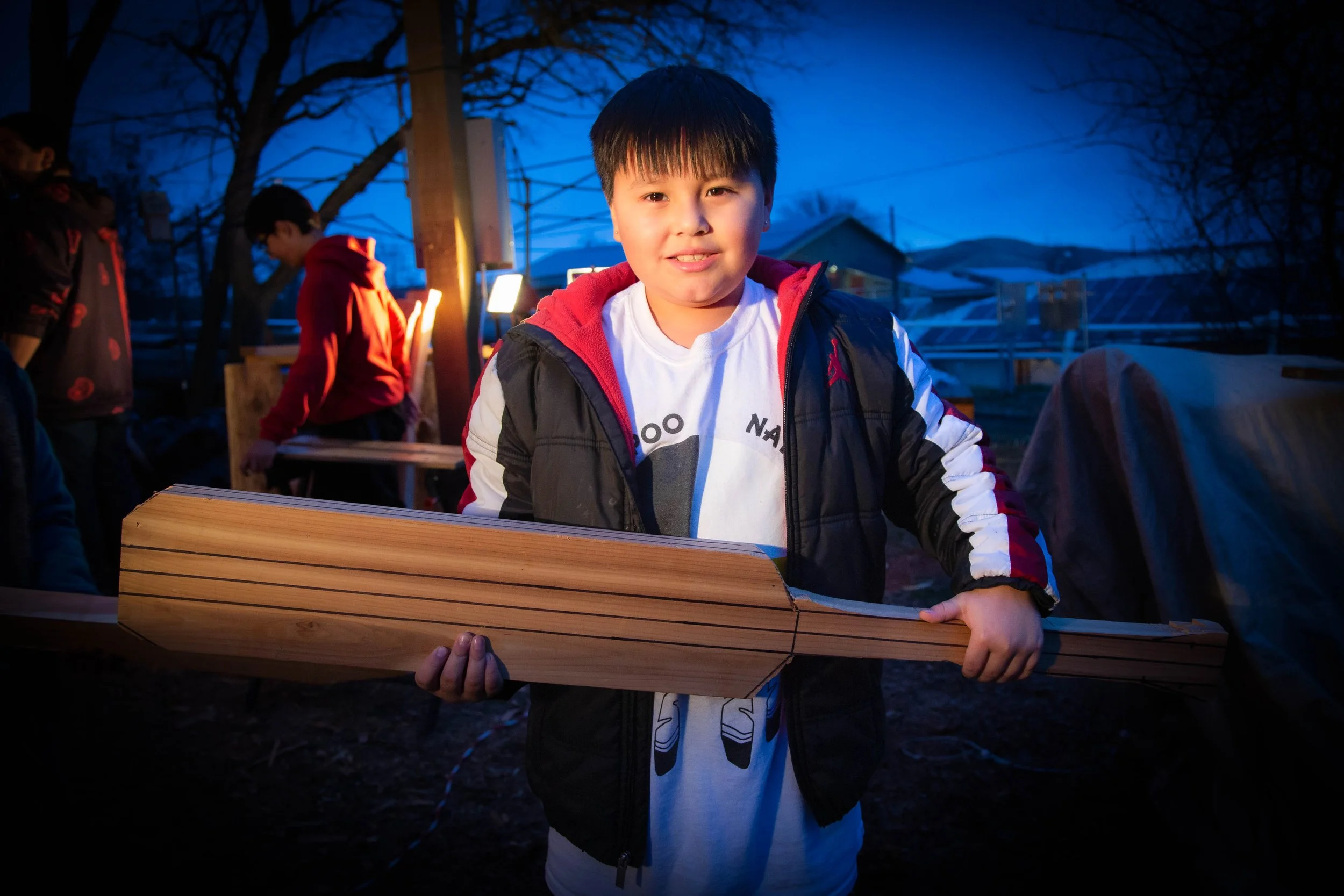 A young boy with dark hair smiling while holding a piece of wood outdoors during dusk. Several other children are in the background, some wearing jackets, with leafless trees and a house behind them.