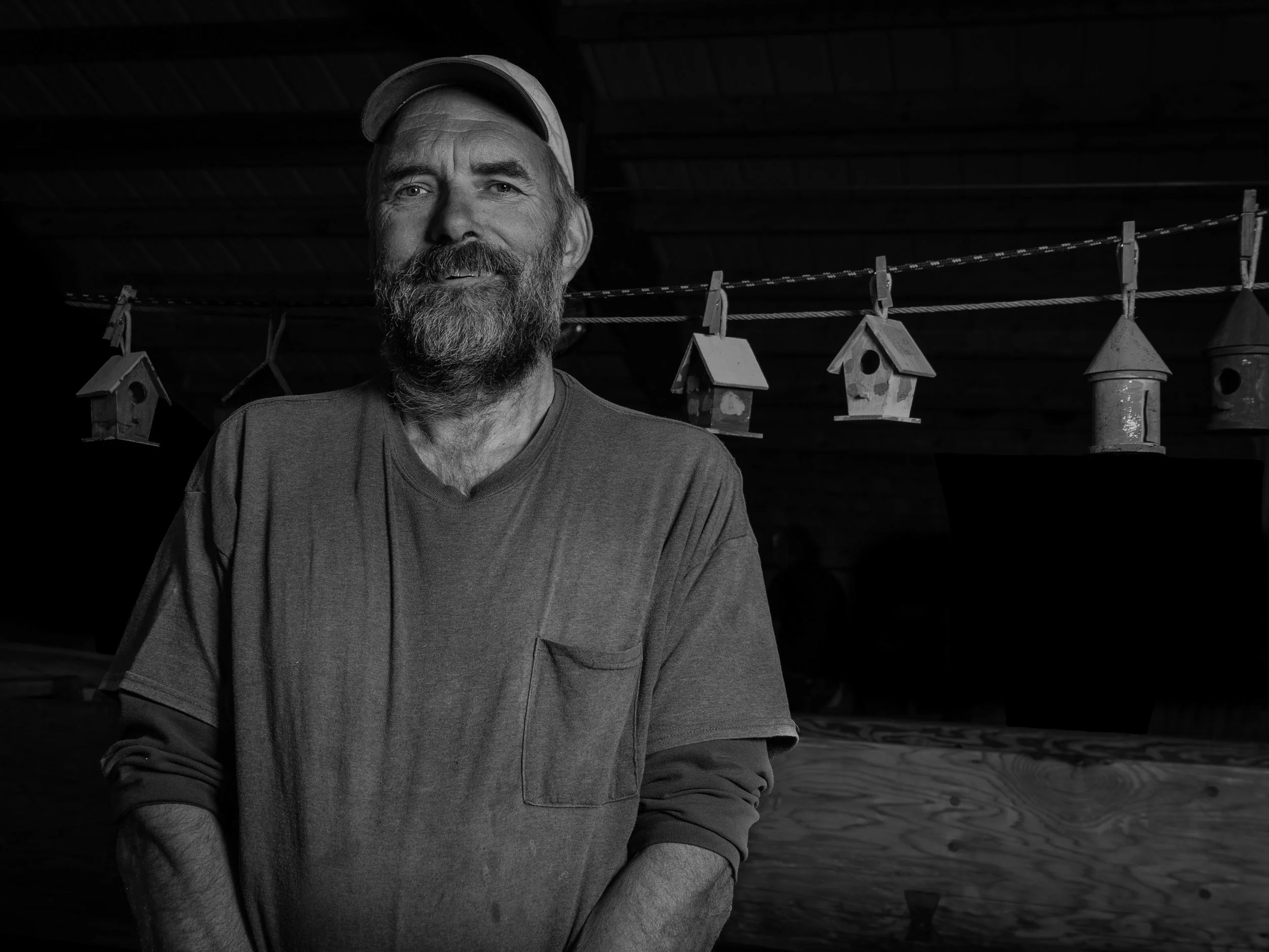 A man with a beard and mustache wearing a baseball cap, standing indoors next to a line of small birdhouses hanging from a string.