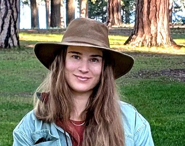 A young woman with long hair wearing a wide-brim hat and denim jacket outdoors in a wooded area with tall trees in the background