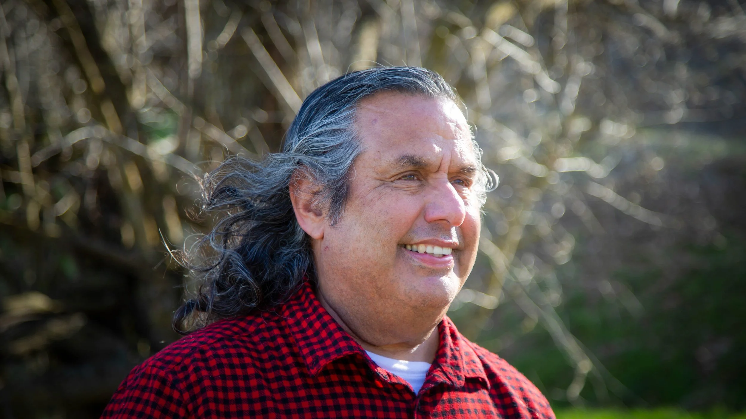 Close-up of a smiling middle-aged man with long gray hair, wearing a red and black checkered shirt, outdoors with blurred trees in the background.