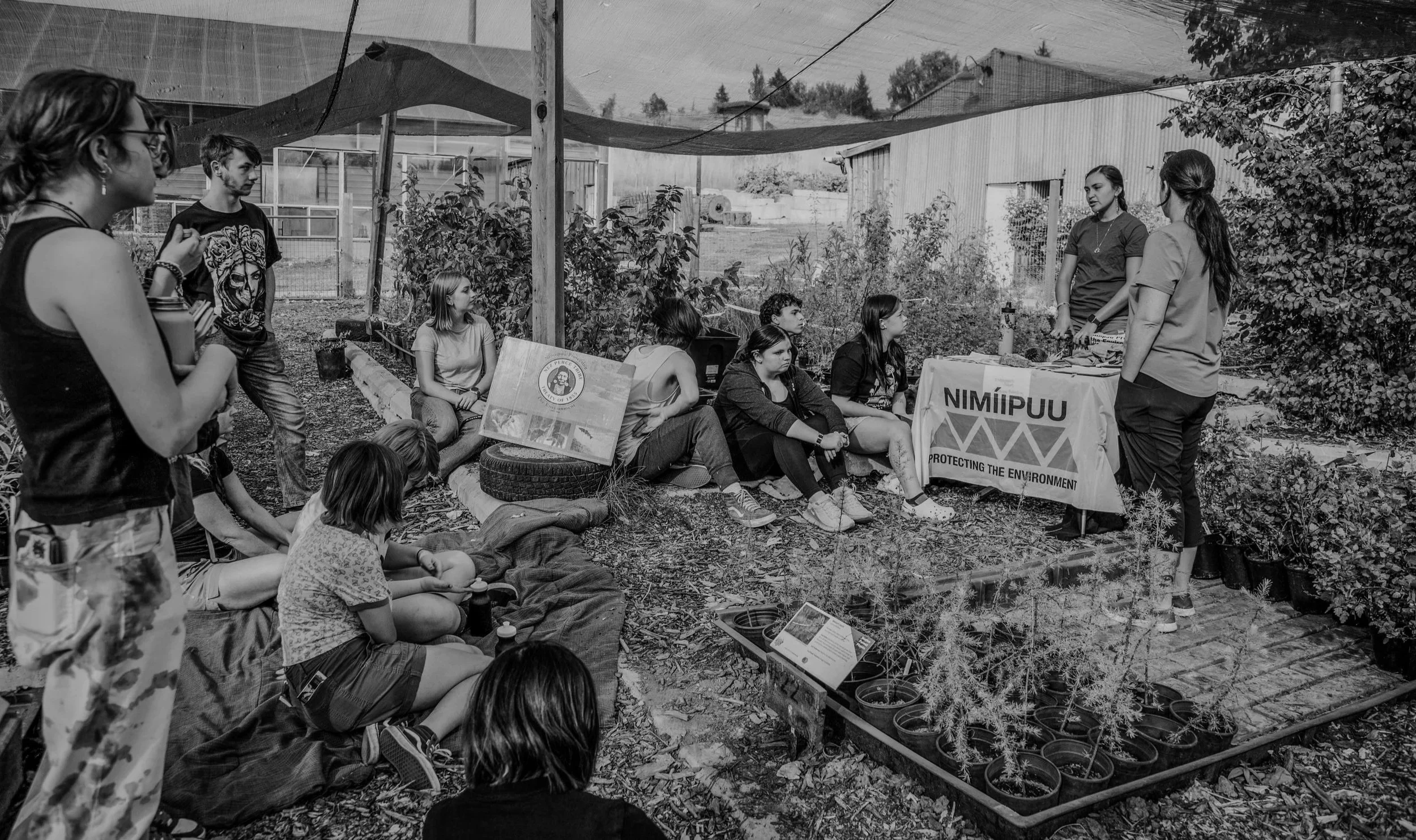 Group of people gathered outdoors under a shade cloth. Some sitting on the ground, others standing, listening to two women speaking. There are plant containers, informational signs, and a banner with the words 'Nimipuu Protecting the Environment'.