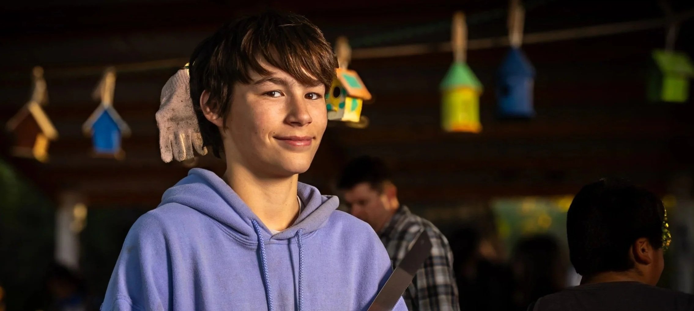 A young man with brown hair and a slight smile, wearing a light purple hoodie, standing indoors at a social gathering with colorful hanging decorations in the background.