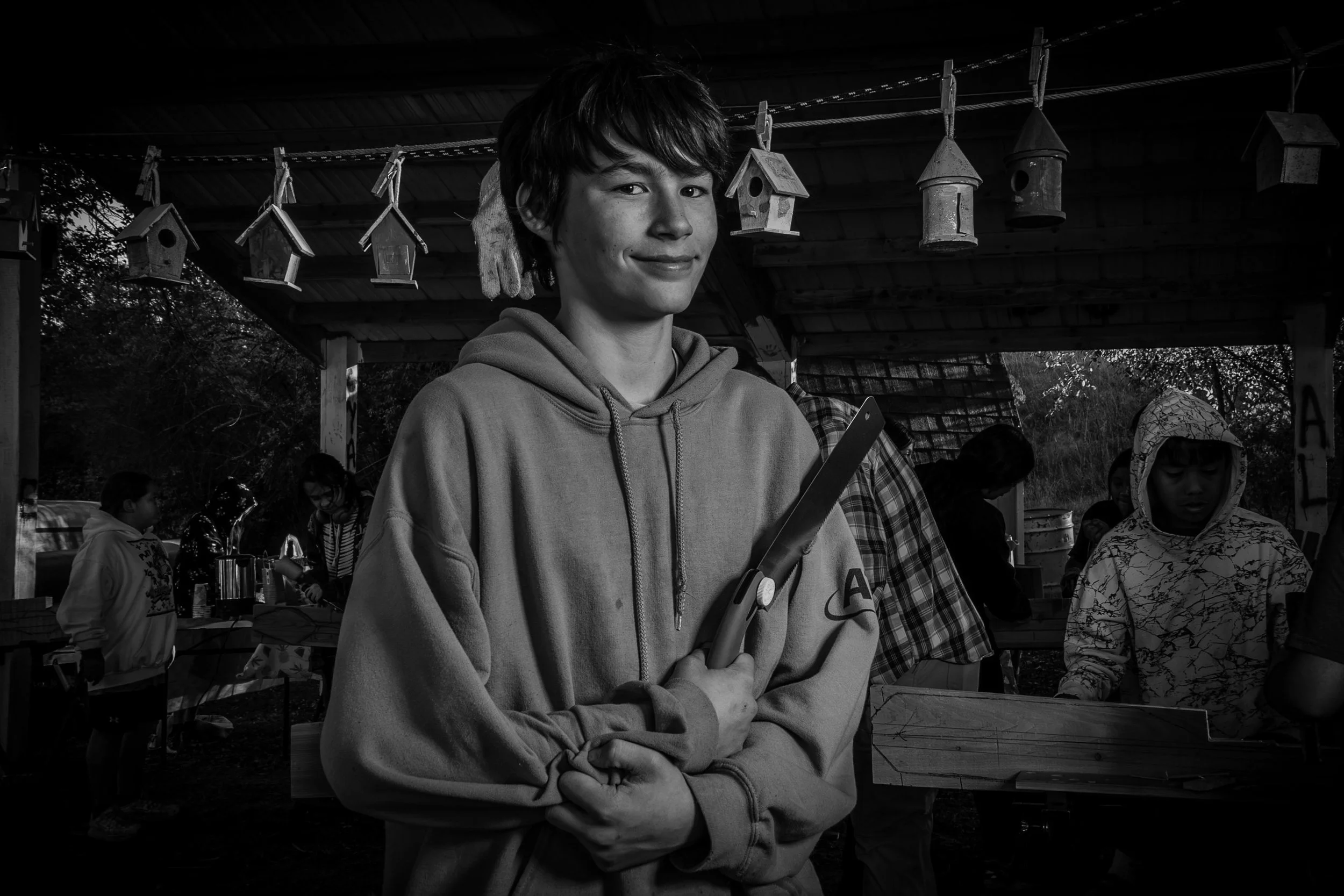 A young man holds a tool while standing outdoors at a gathering or event with other people, some of whom are wearing hoodies and jackets, in a setting with birdhouses hanging from a line in the background.
