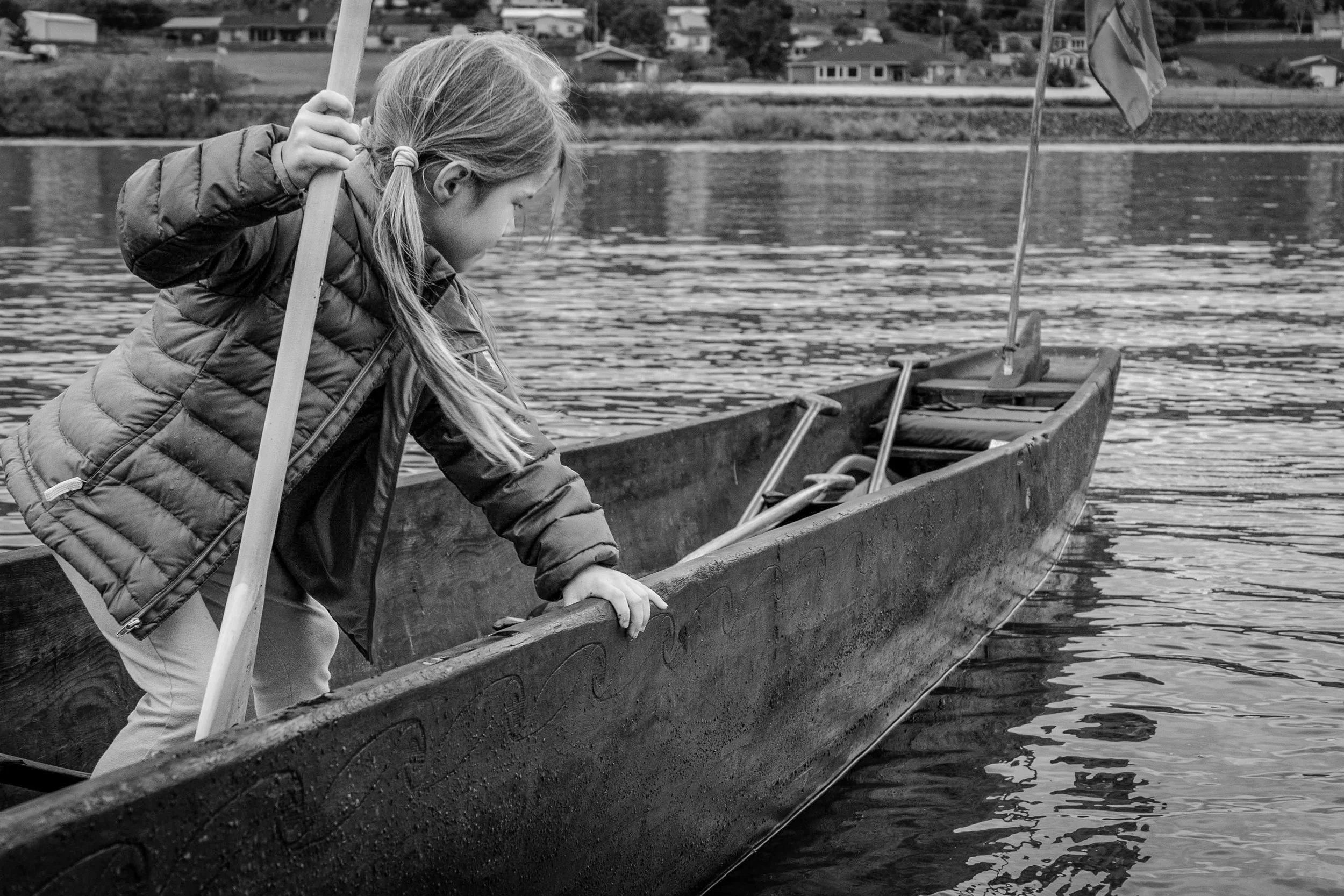 A girl with long hair tied in a ponytail, wearing a puffy jacket, is leaning over the side of a canoe on a lake, holding a wooden paddle. The water is calm with ripples, and in the background, there are houses and trees along the shoreline.