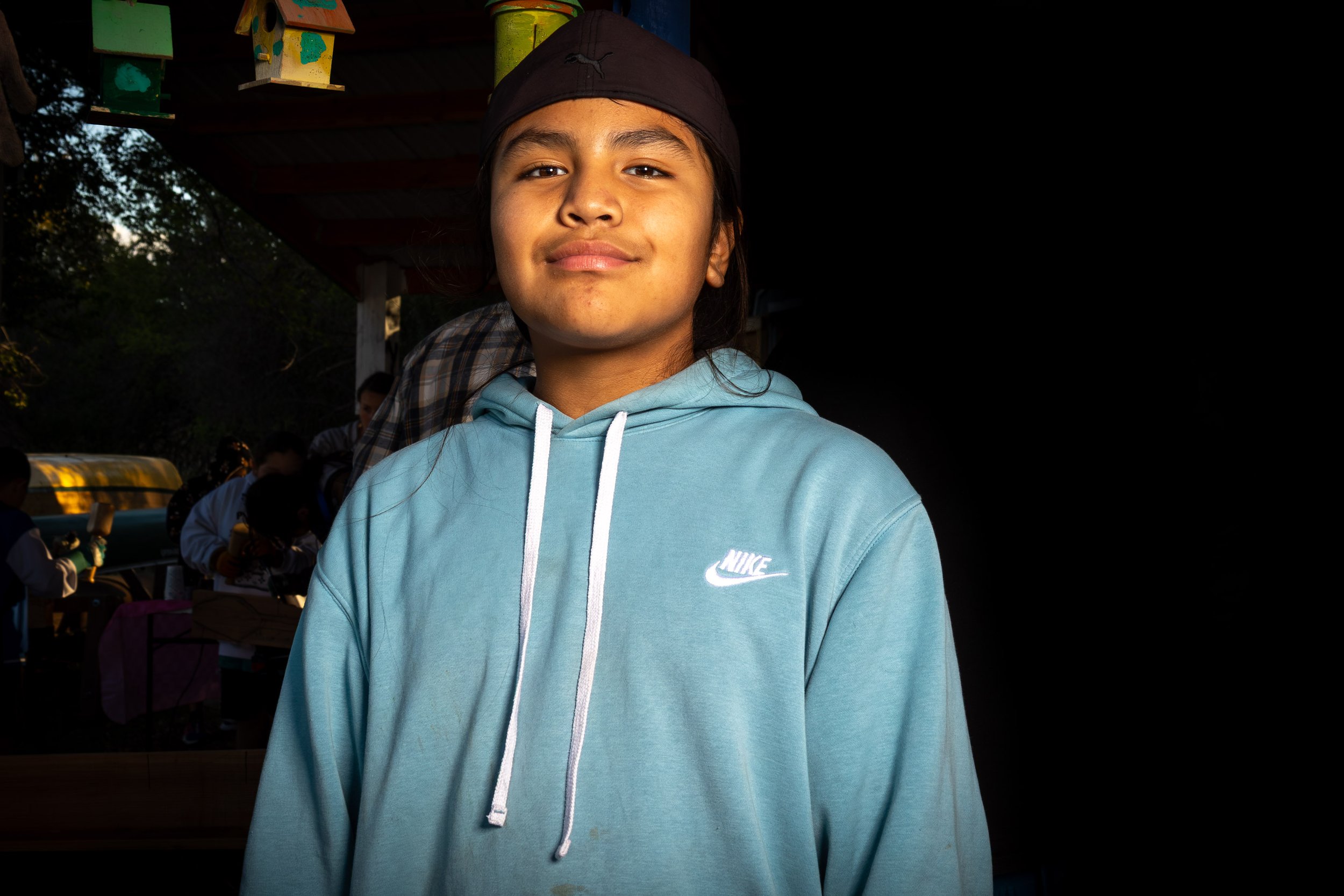 A young person with medium skin tone wearing a black head covering and light blue Nike hoodie, smiling slightly, standing outdoors with trees and a wooden structure in the background.