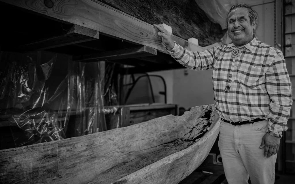 A man smiling and wearing a checkered shirt, standing in a workshop with a large wooden boat. He has his hand on the boat, which is under construction or restoration.