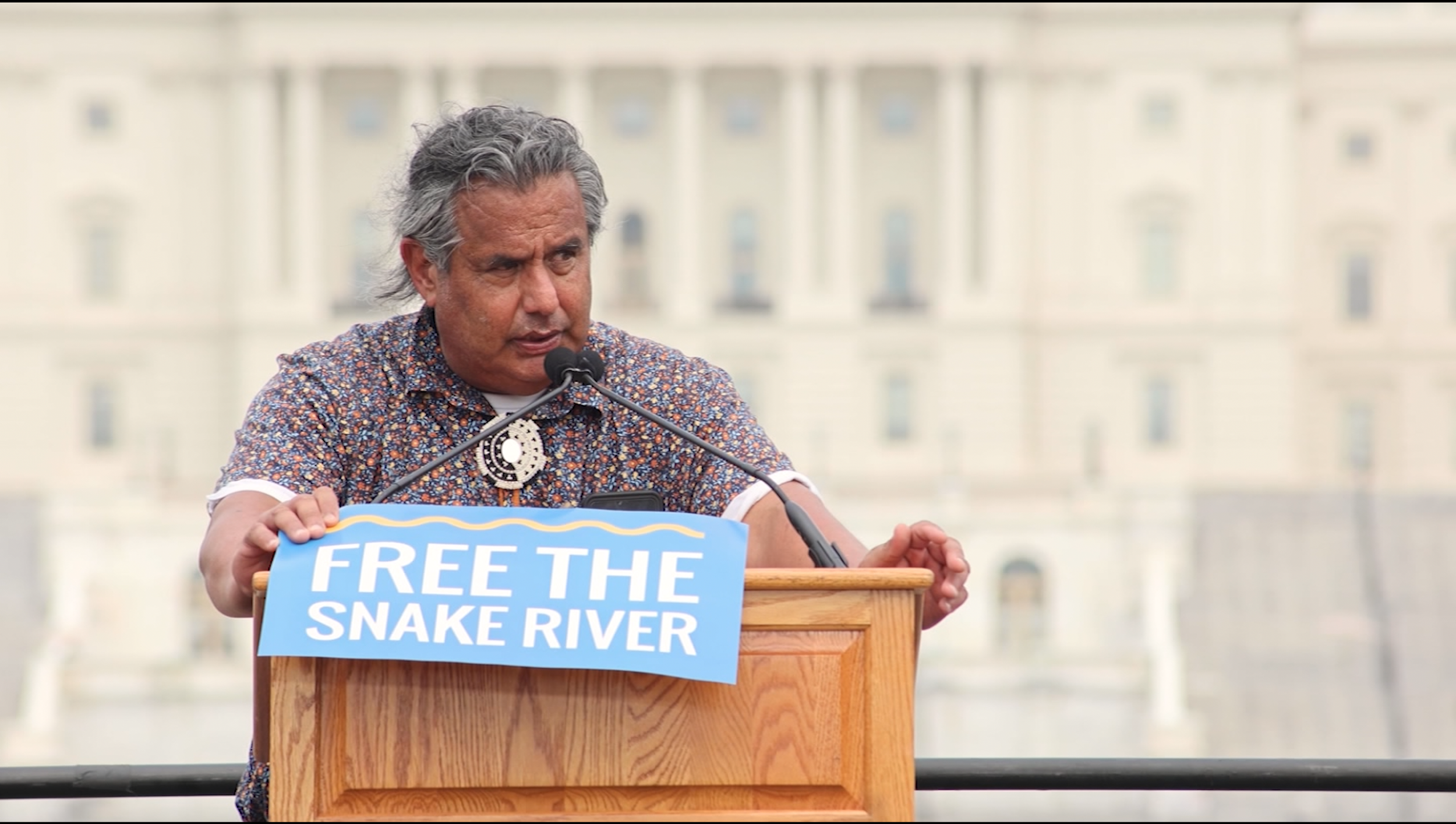 A man with gray hair speaking at a podium outdoors, holding a blue sign that reads 'FREE THE SNAKE RIVER.' In the background, there is a large government building.