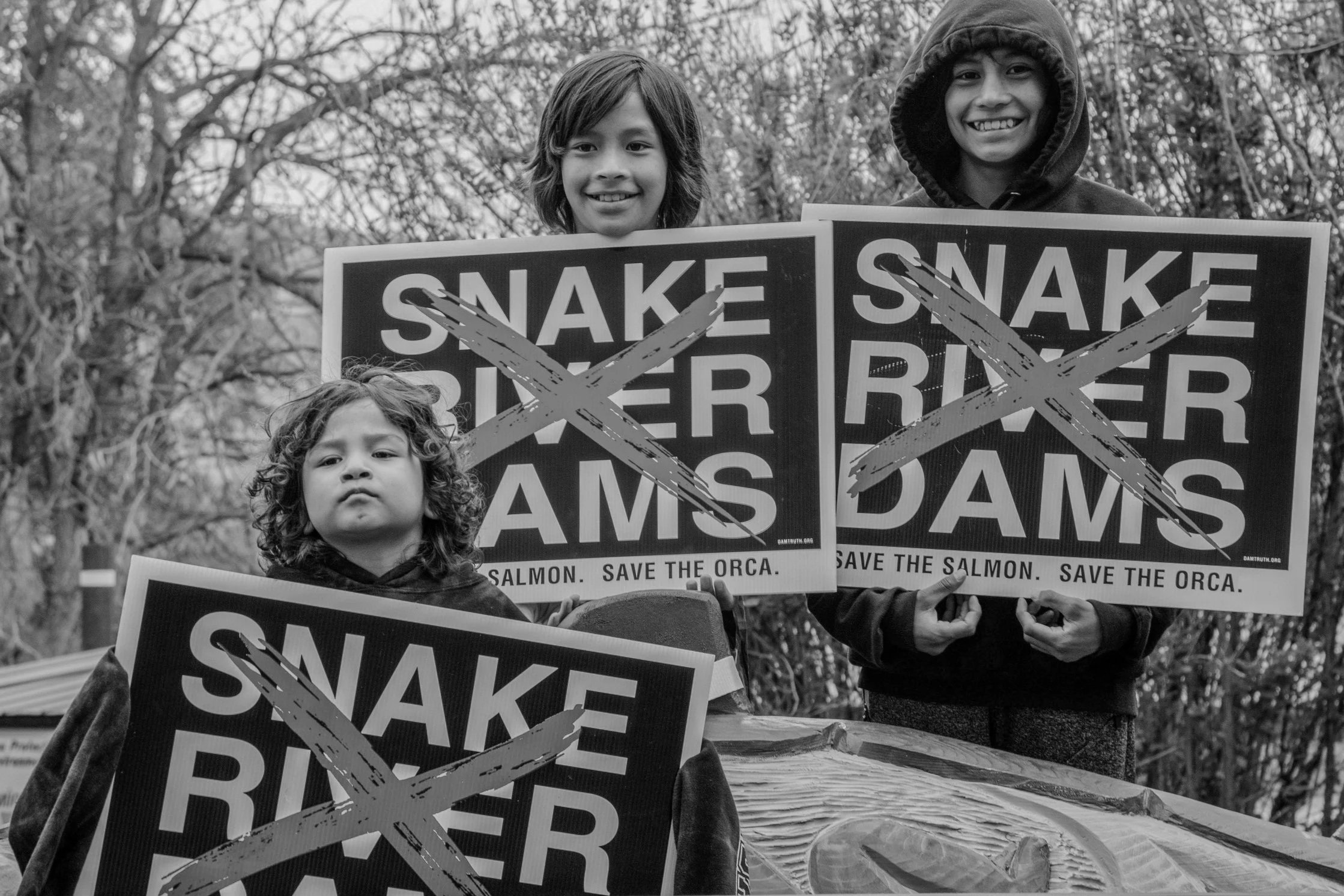 Black and white photo of three children holding protest signs that say 'Save the Salmon' and 'Save the Orca'. The signs have large black X marks across them. The children are outdoors, with trees in the background.