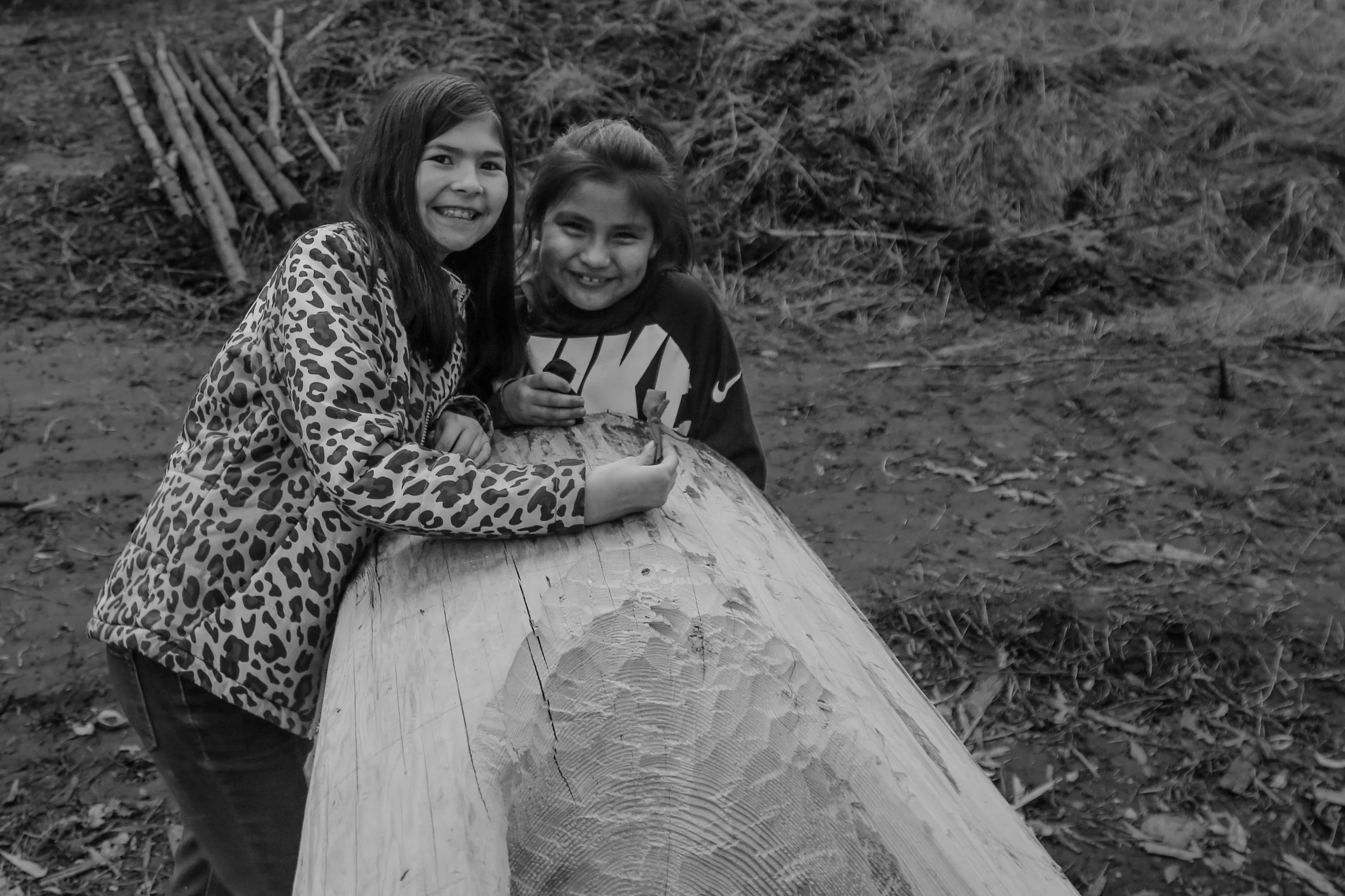 Two young girls smiling while leaning on a large cut log outdoors, with a backdrop of bare ground and scattered logs.