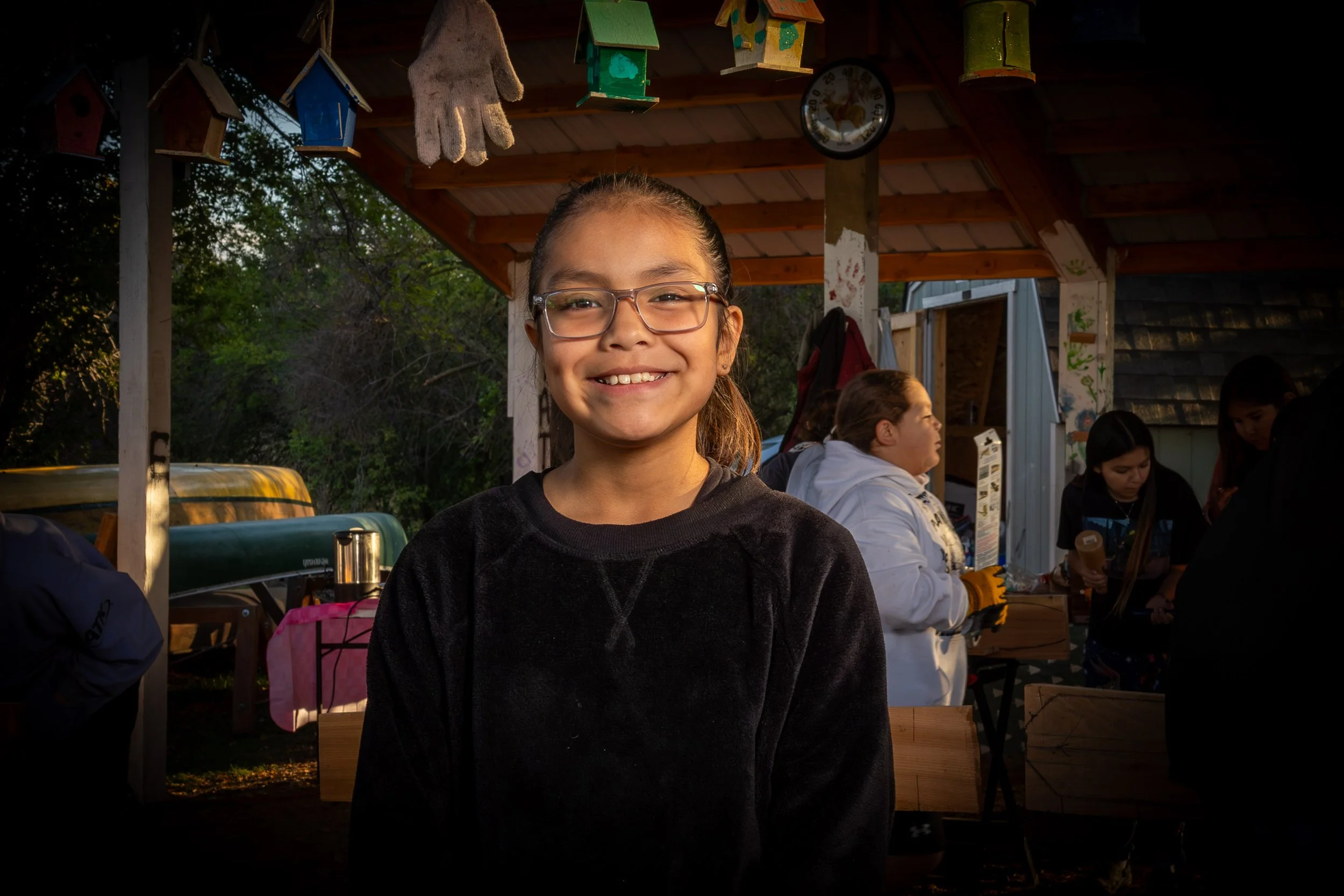 Smiling girl wearing glasses standing in an outdoor area under a wooden roof with children and adults in the background, hanging birdhouses above.