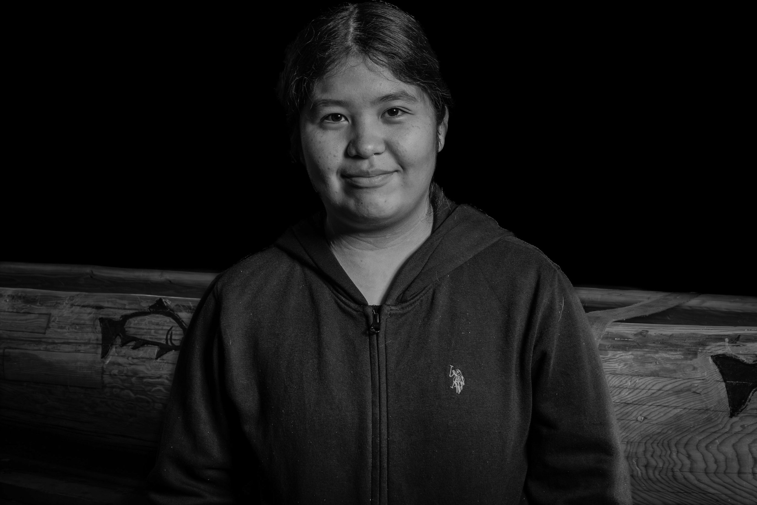 Black and white portrait of a woman with dark hair, wearing a hoodie, standing outdoors in front of a wooden fence.