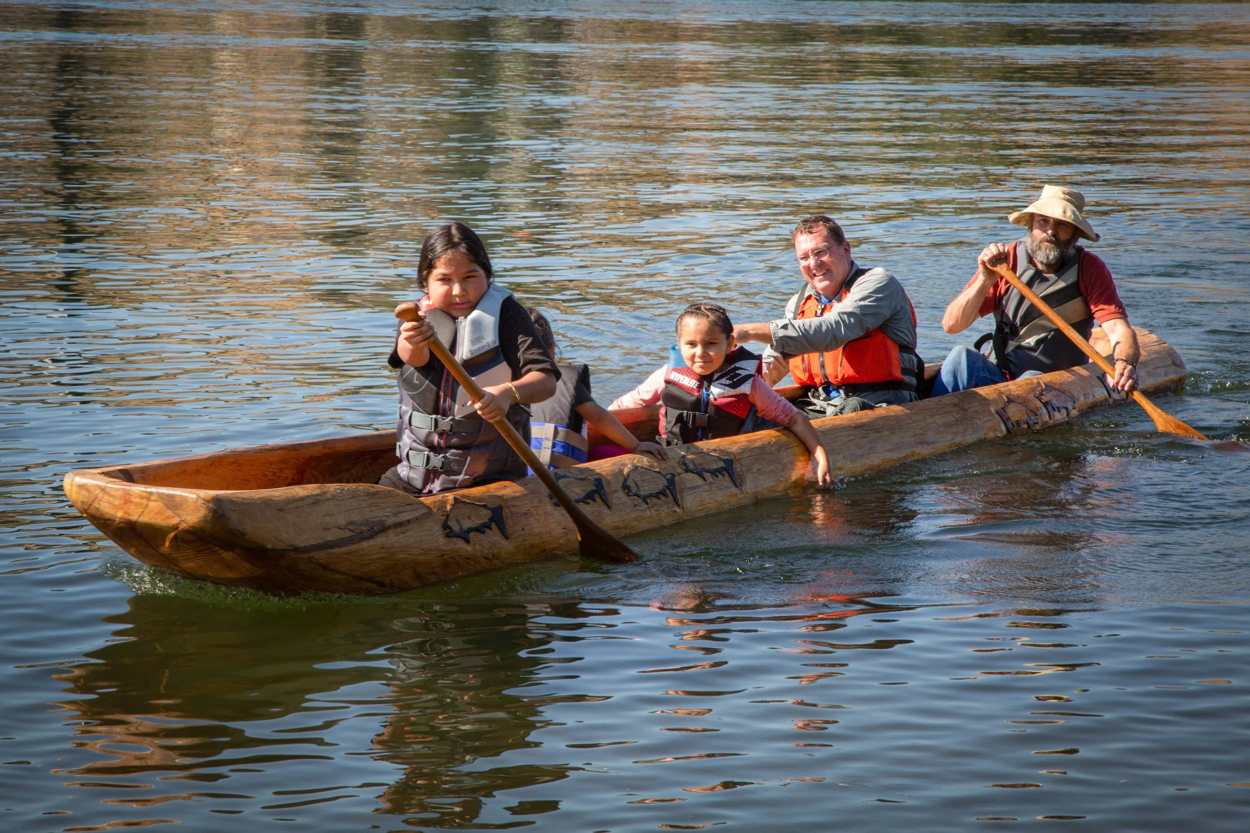 Group of four people, two adults and two children, paddling a wooden canoe on a body of water, with trees reflected in the water.
