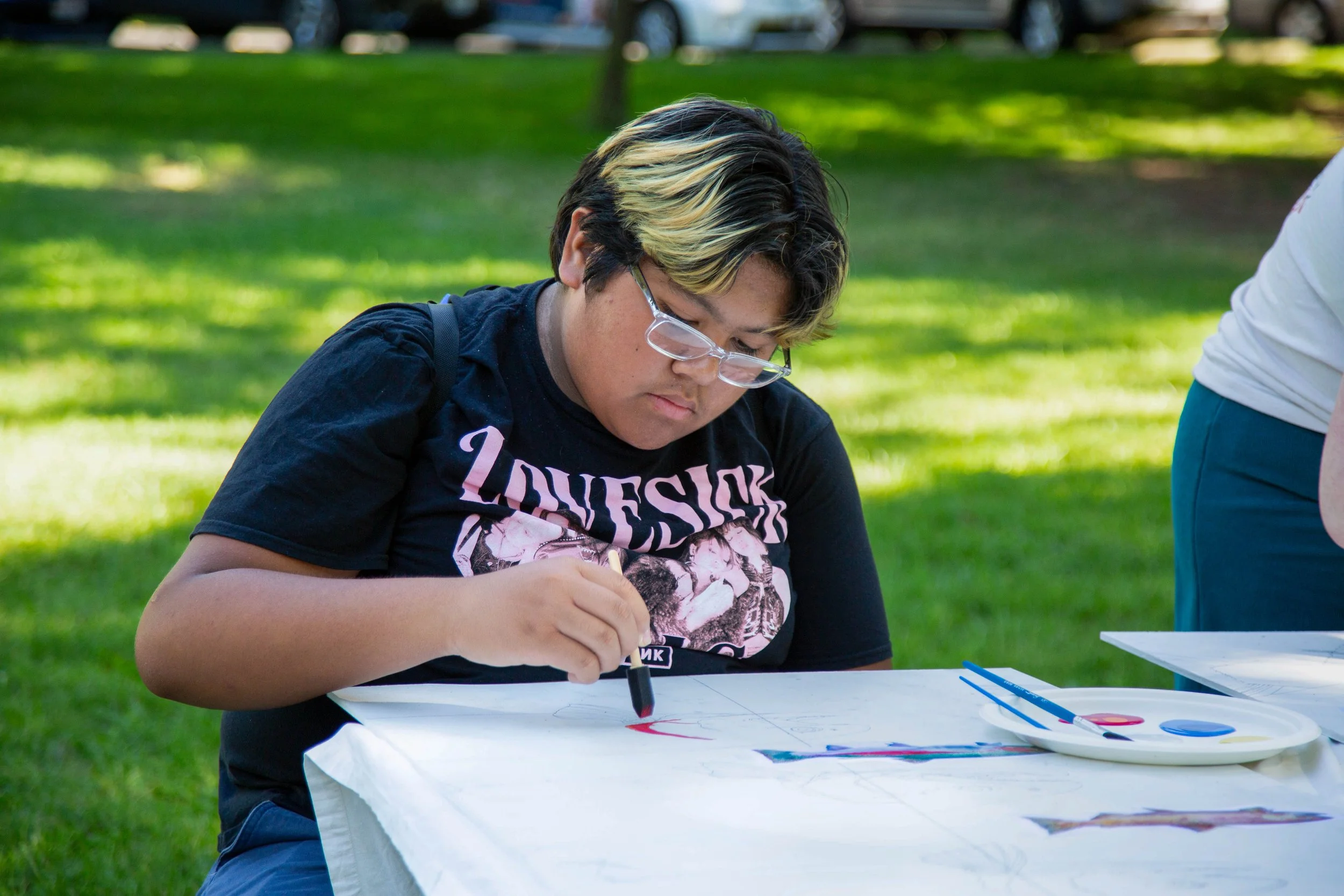 A young person with glasses and black hair with blonde streaks, sitting at a table outdoors, drawing with colored markers on paper, with green grass and trees in the background.