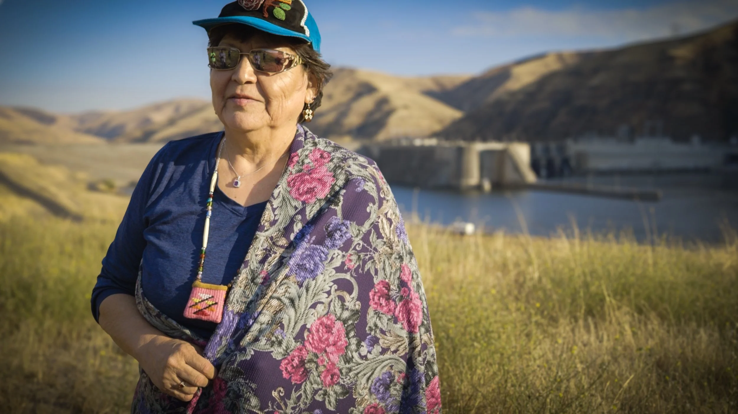 An elderly woman wearing sunglasses, a blue cap, and floral jacket standing outdoors with a dam and hills in the background.