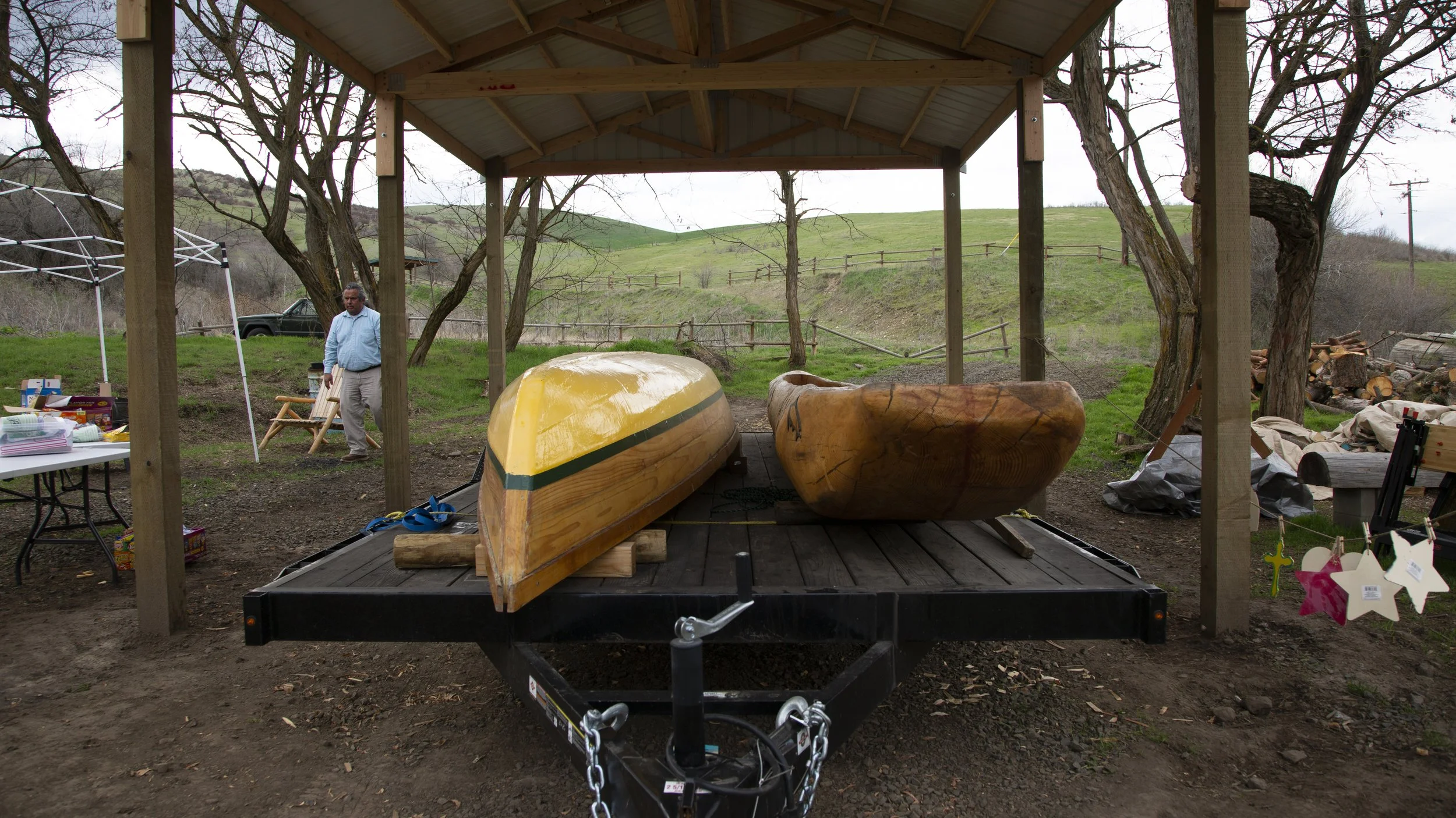 A wooden boat on a trailer under a covered outdoor area, with a man in a blue shirt standing nearby. The area has trees, green rolling hills, and various objects and supplies on tables and the ground.