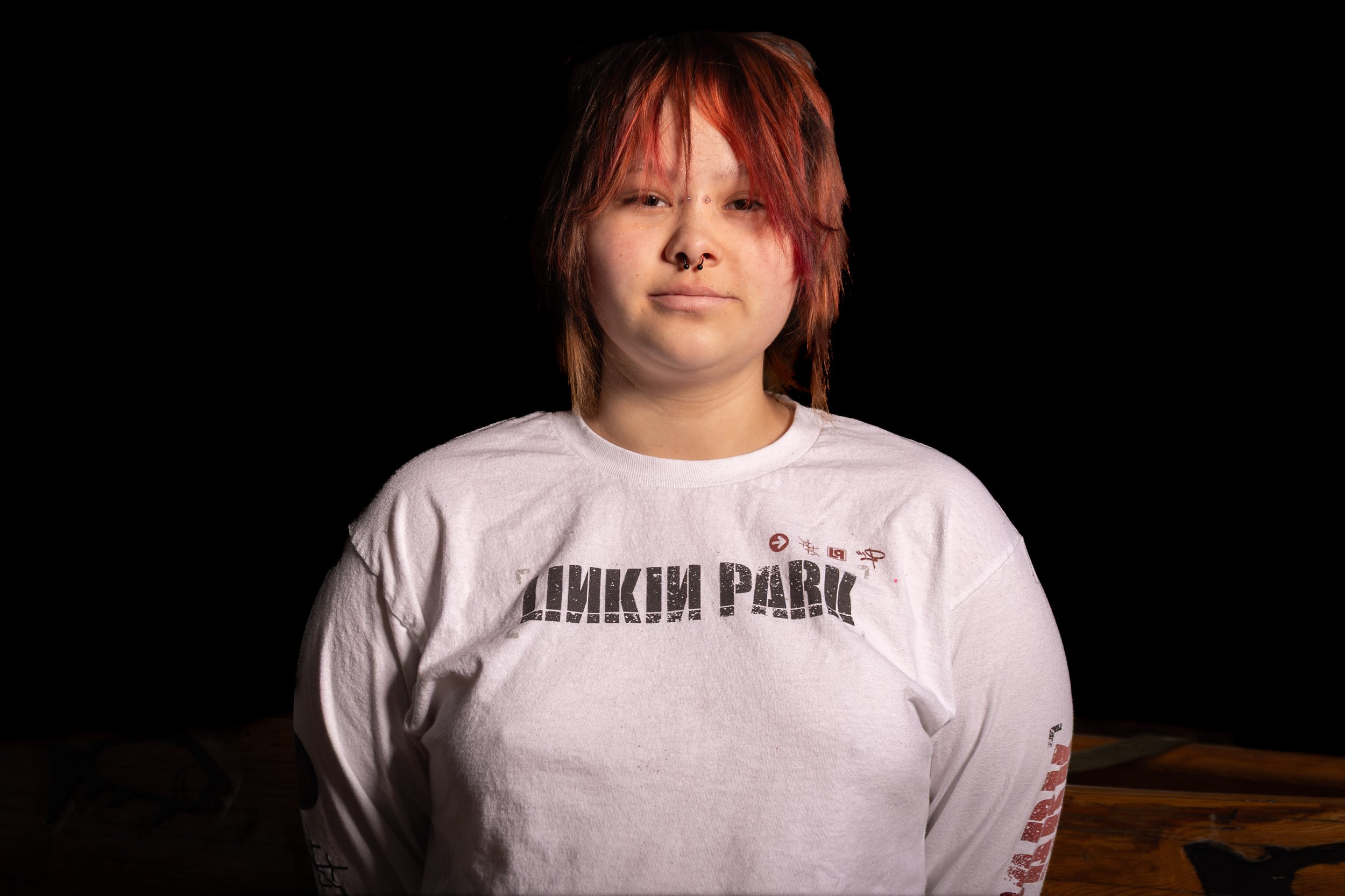 A person with dyed red hair and facial piercings wearing a white Nirvana t-shirt, standing against a black background.