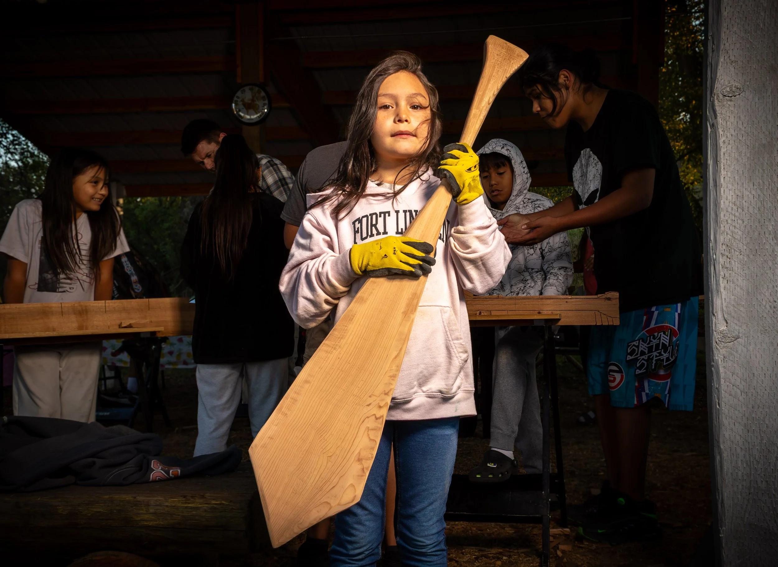 A young girl with long brown hair and yellow gloves holding a wooden paddle, standing in front of other children and adults under a pavilion.