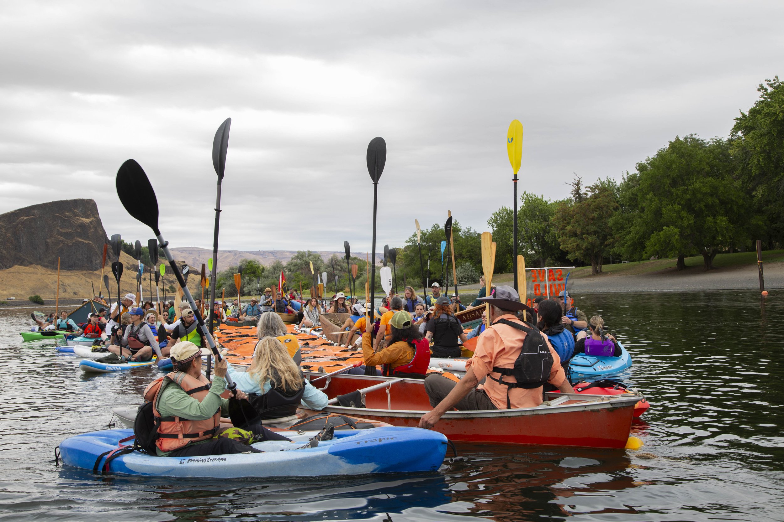 People kayaking and paddleboarding on a river during an outdoor event with a crowd gathered on the water and on land, under a cloudy sky with green trees and a rocky landscape in the background.