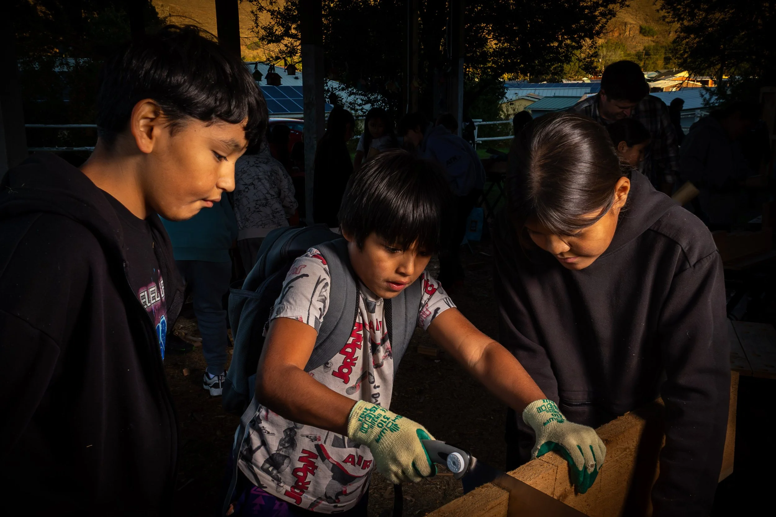 Children and adults working on a woodworking project outdoors at sunset, wearing gloves and focused on their tasks.