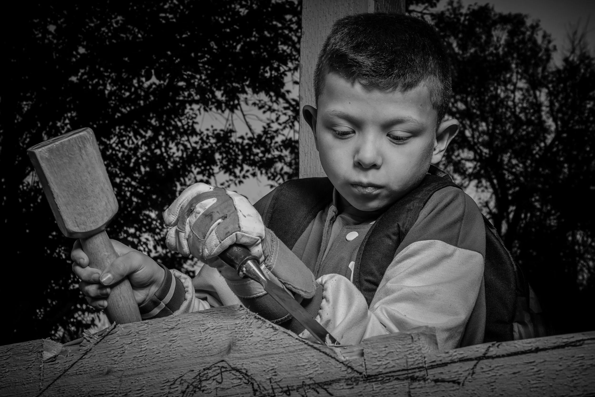 Black and white photo of a young boy holding a hammer and a saw, working on a piece of wood outdoors.