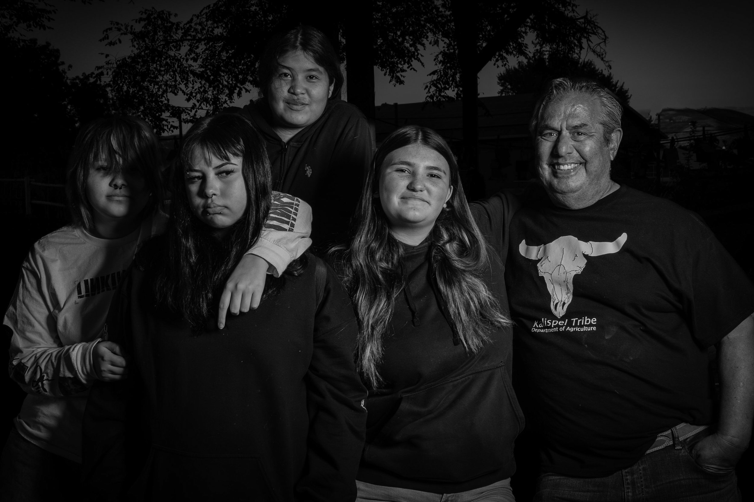 A group photo of five people, including children and an adult, smiling and posing outdoors at dusk or night. The background shows trees and a building, with some lighting.