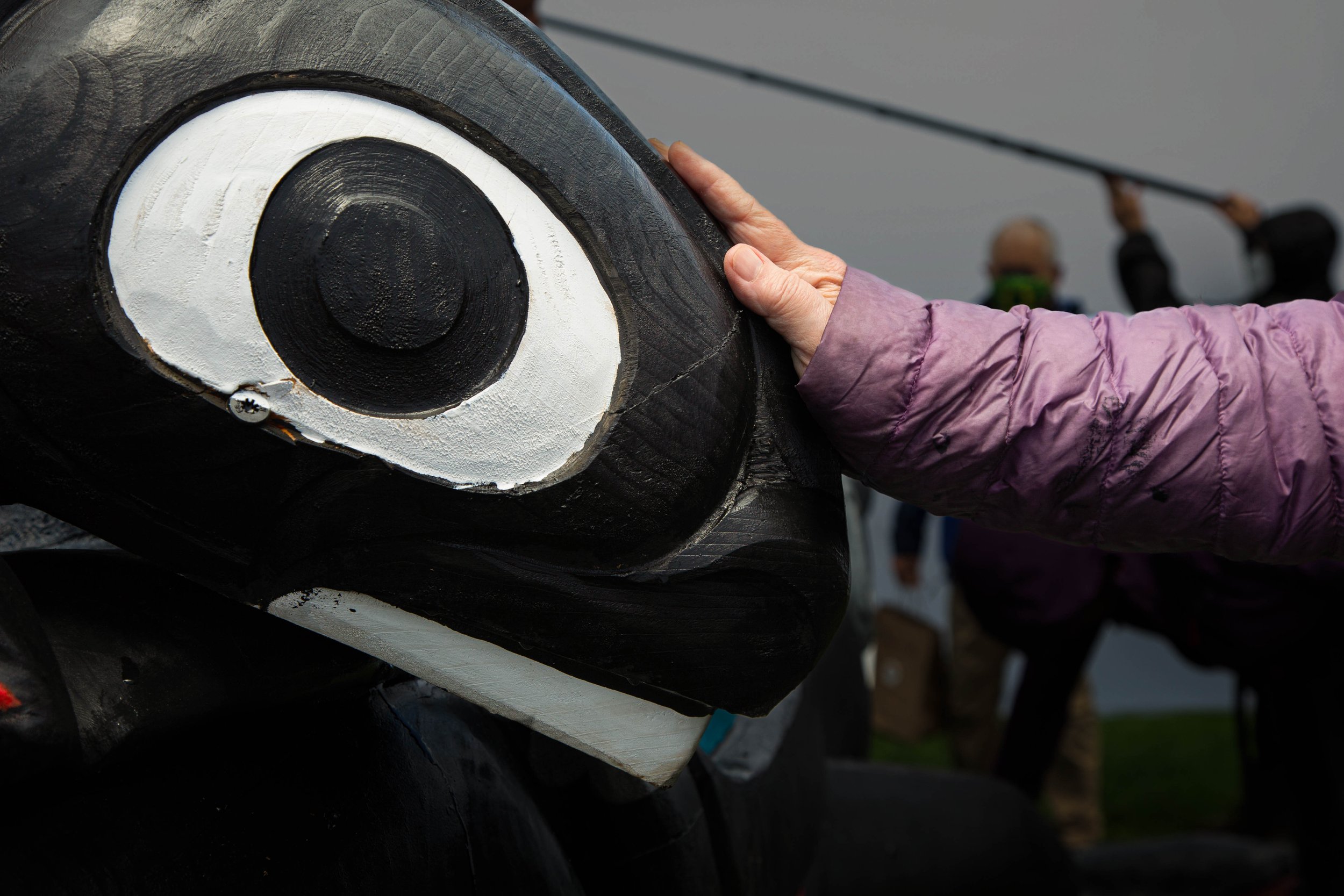 A person wearing a purple jacket touches the eye painted on a black wooden or carved statue, which has white and black colors for the eye, with a background of out-of-focus individuals and some circus or festival lights.