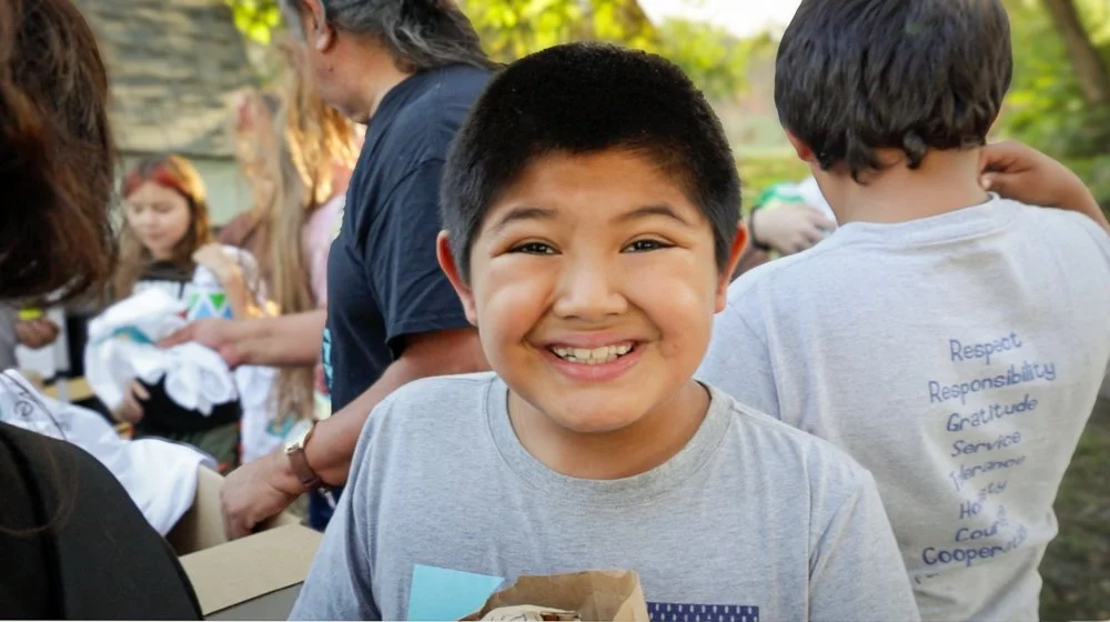 Smiling boy with short dark hair in gray shirt standing outdoors, surrounded by children and adults.