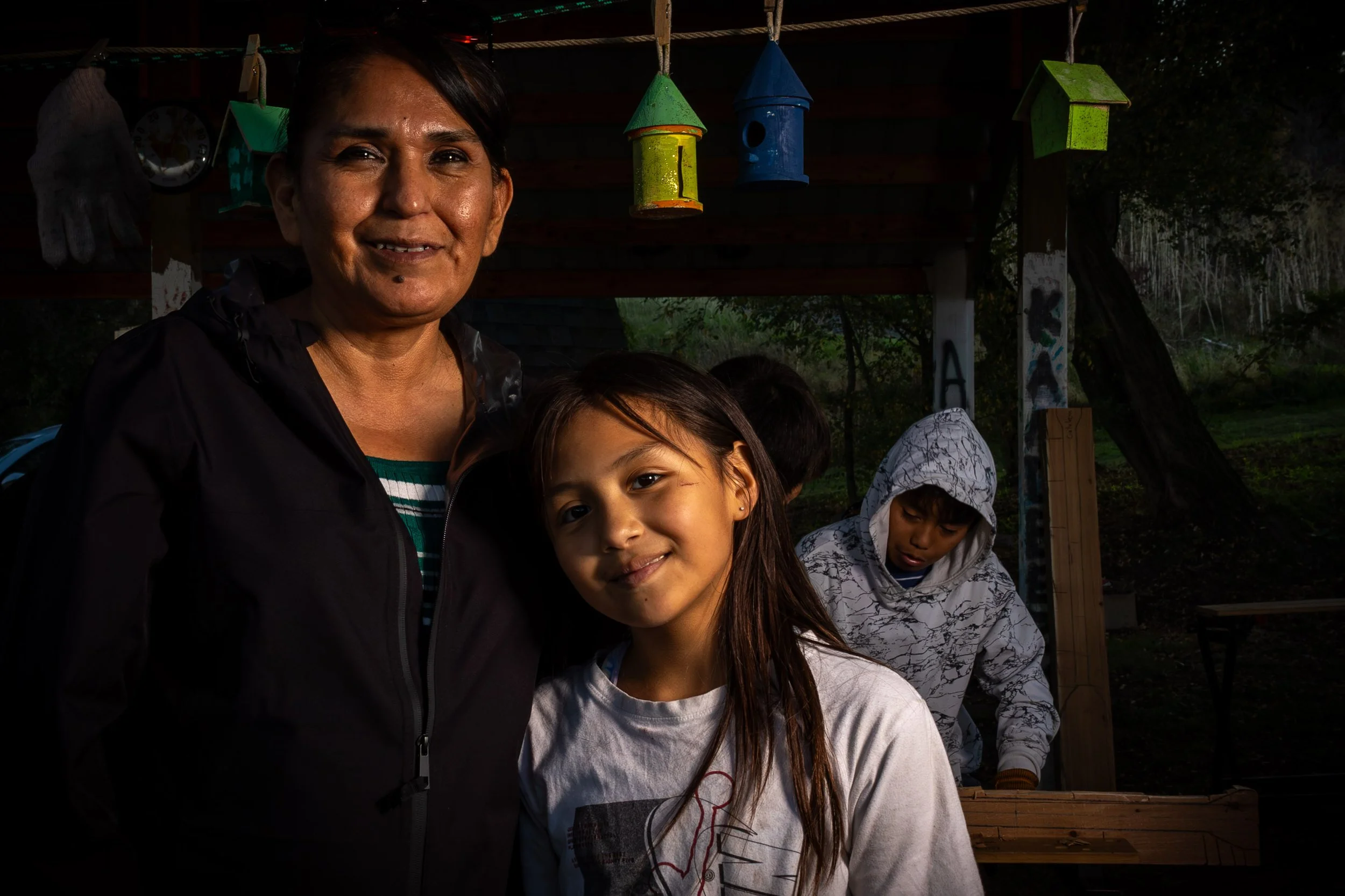 A woman and a young girl are smiling in an outdoor setting at dusk or early evening. There are colorful birdhouses hanging above and children in the background. The scene includes natural foliage and a wooden structure.