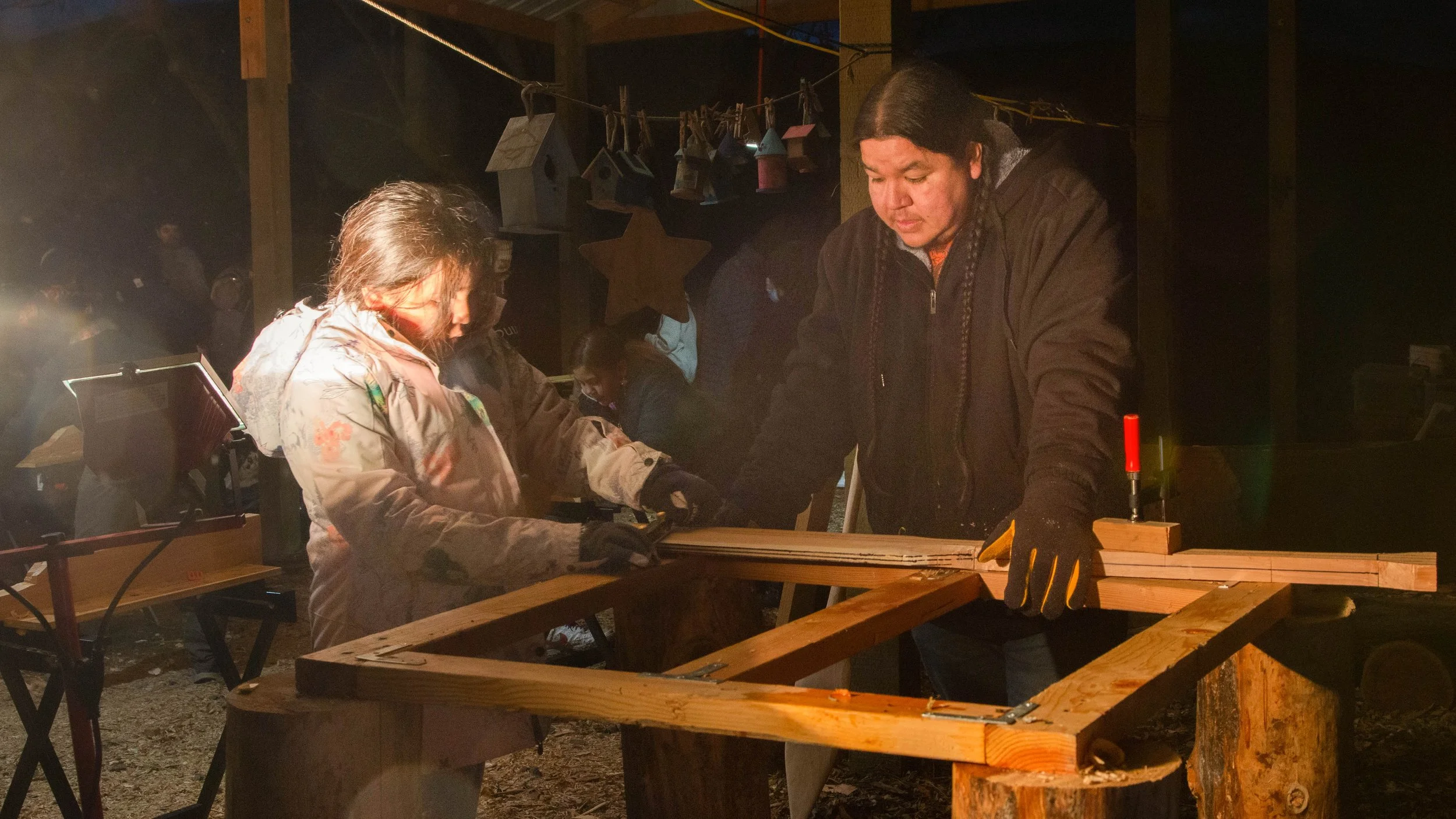 Two people working together on woodworking in a dimly lit workshop or cabin. One person is an adult man and the other is a young girl. They are focused on a wooden project, surrounded by tools and supplies. Birdhouses are hanging in the background.