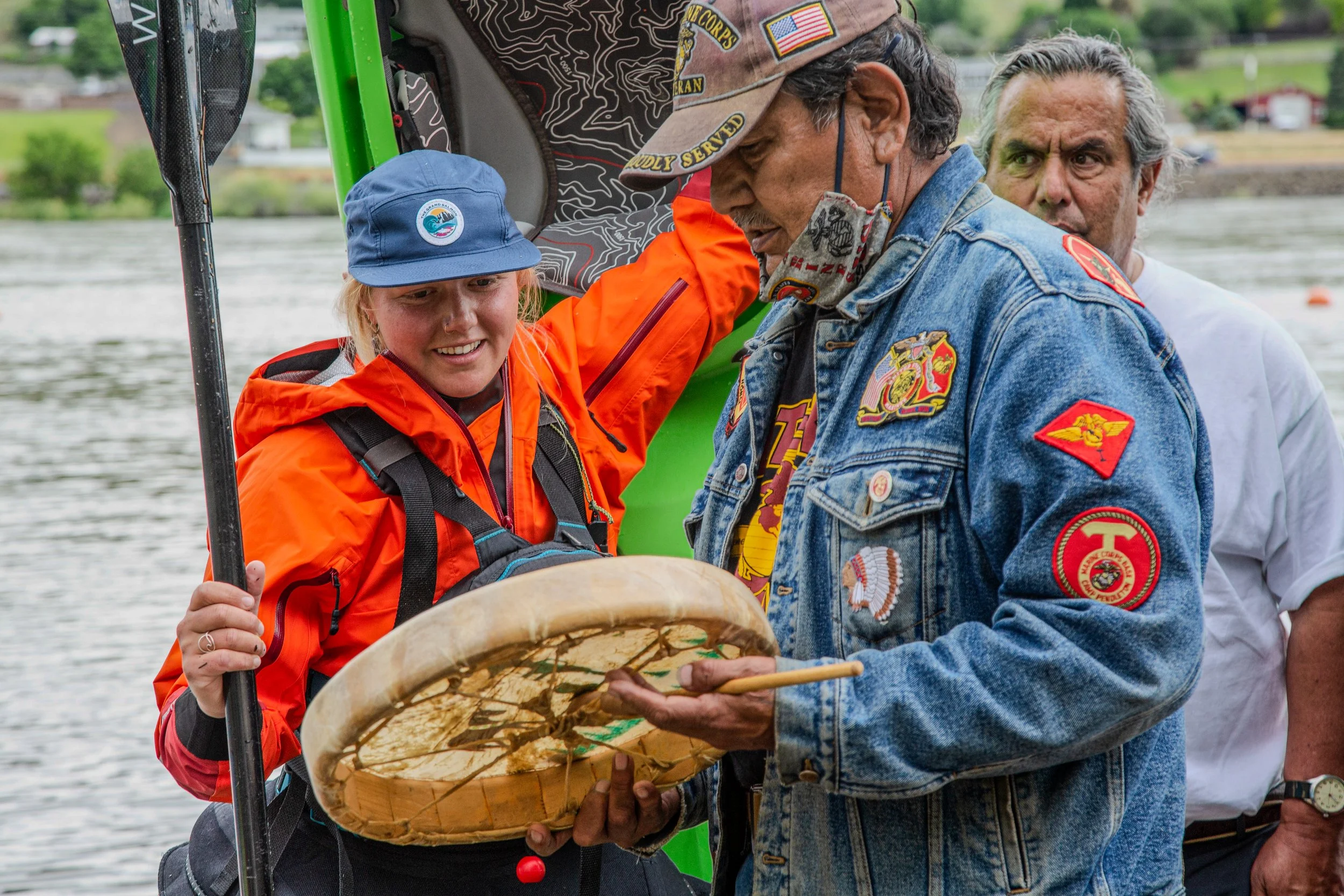 A young woman wearing an orange jacket and a blue hat with a logo, holding a paddle, is looking at an older man with a patch on his denim jacket and a cap with military patches, while he holds a basket with hands inside it during an outdoor event by 