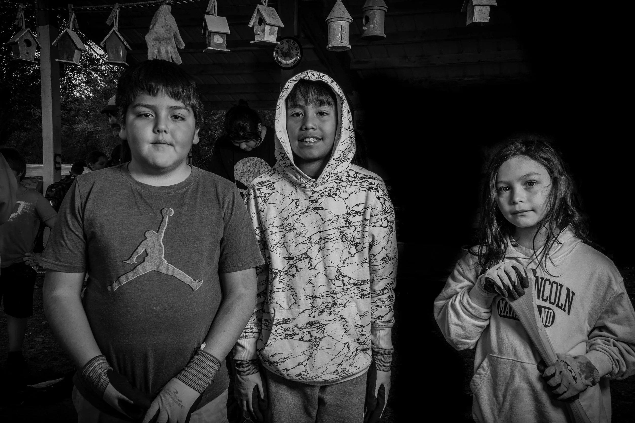 A black-and-white photo of three children standing outdoors, two boys in the foreground, and a girl to the right. The boy on the left has short hair and wears a t-shirt with a basketball player logo. The boy in the middle has a hoodie with a marbled pattern and is smiling slightly. The girl has long wavy hair, wears a sweatshirt with text, and is holding gloves. There are children and hanging birdhouses in the background.