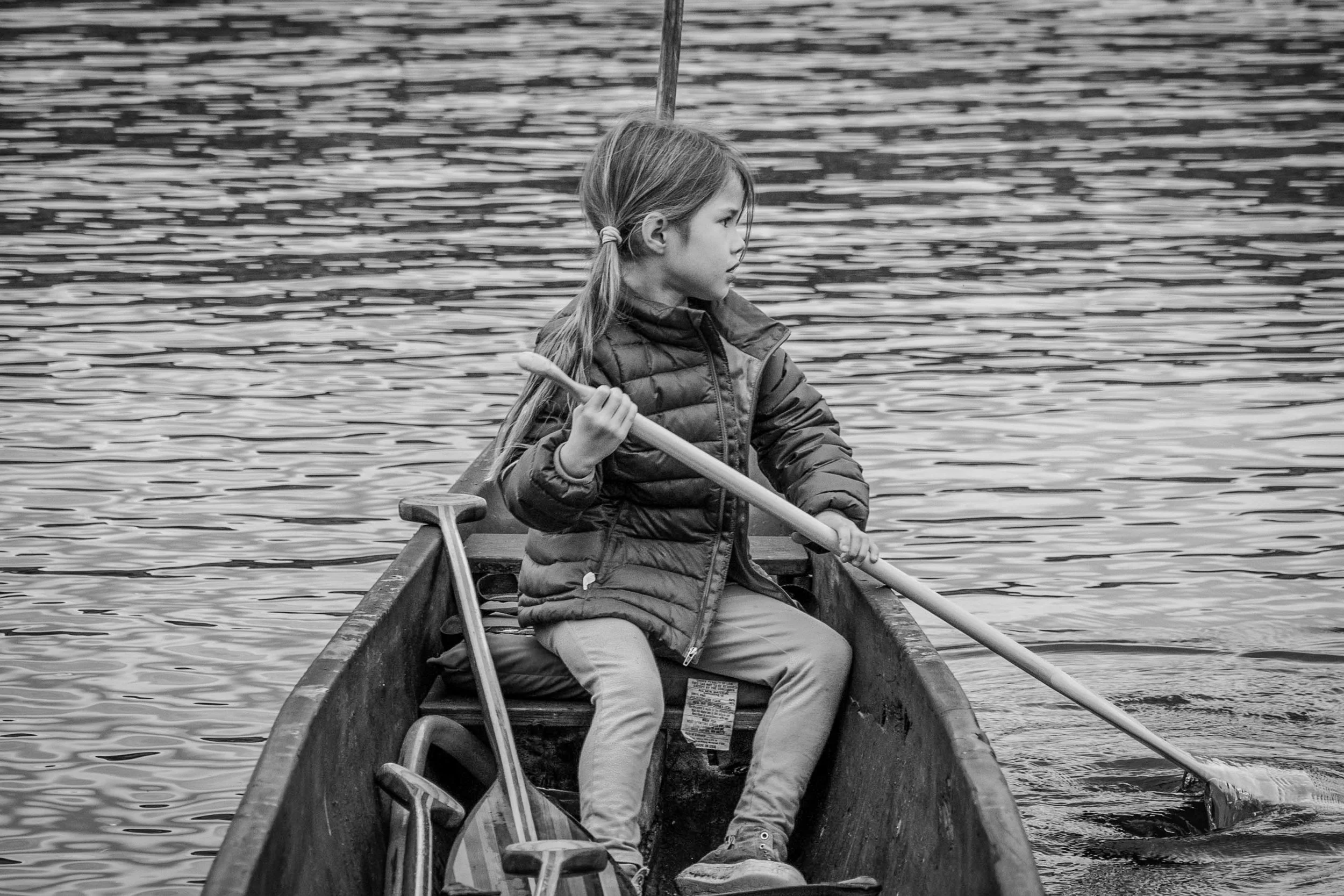 A young girl with long hair in pigtails, wearing a quilted jacket and pants, sitting in a canoe on a body of water, holding a paddle. The water has ripples and appears calm.
