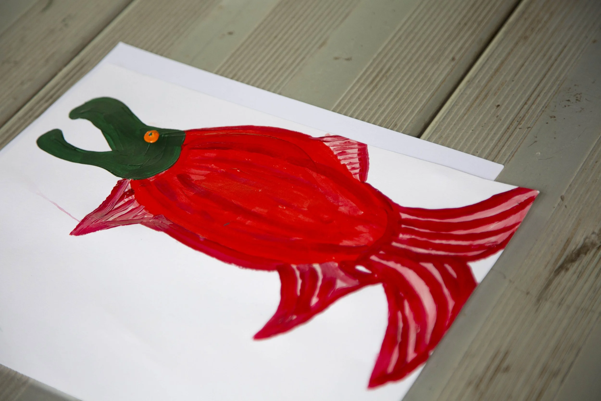 Child's drawing of a dinosaur painted with red, green, and orange colors on white paper placed on a wooden surface.