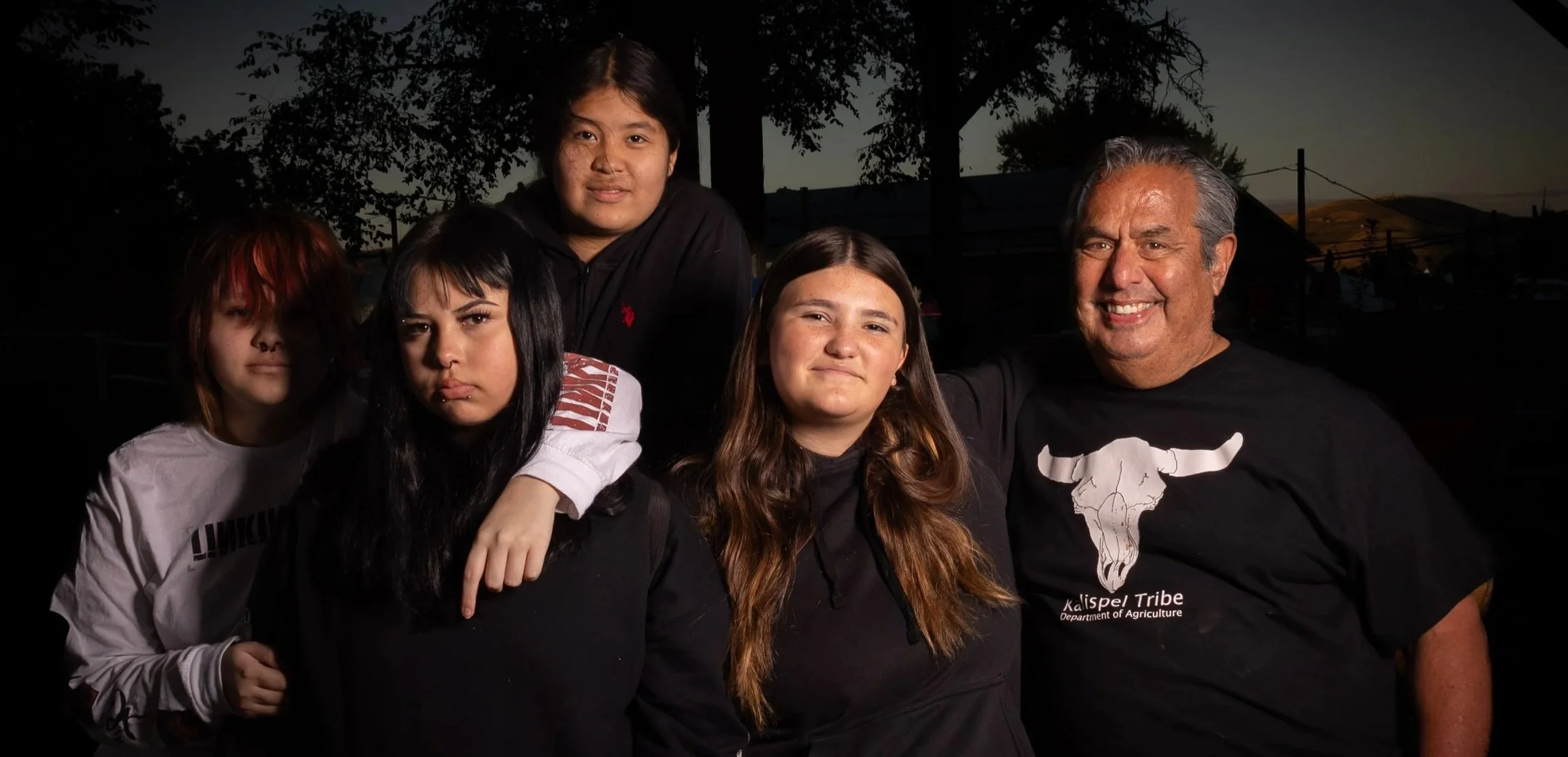 Group of five people, including children and an adult, posing outdoors at dusk. The adult male is wearing a black t-shirt with a white longhorn logo and the words 'Kalsipel Tribe Department of Agriculture.' The children have diverse appearances and a