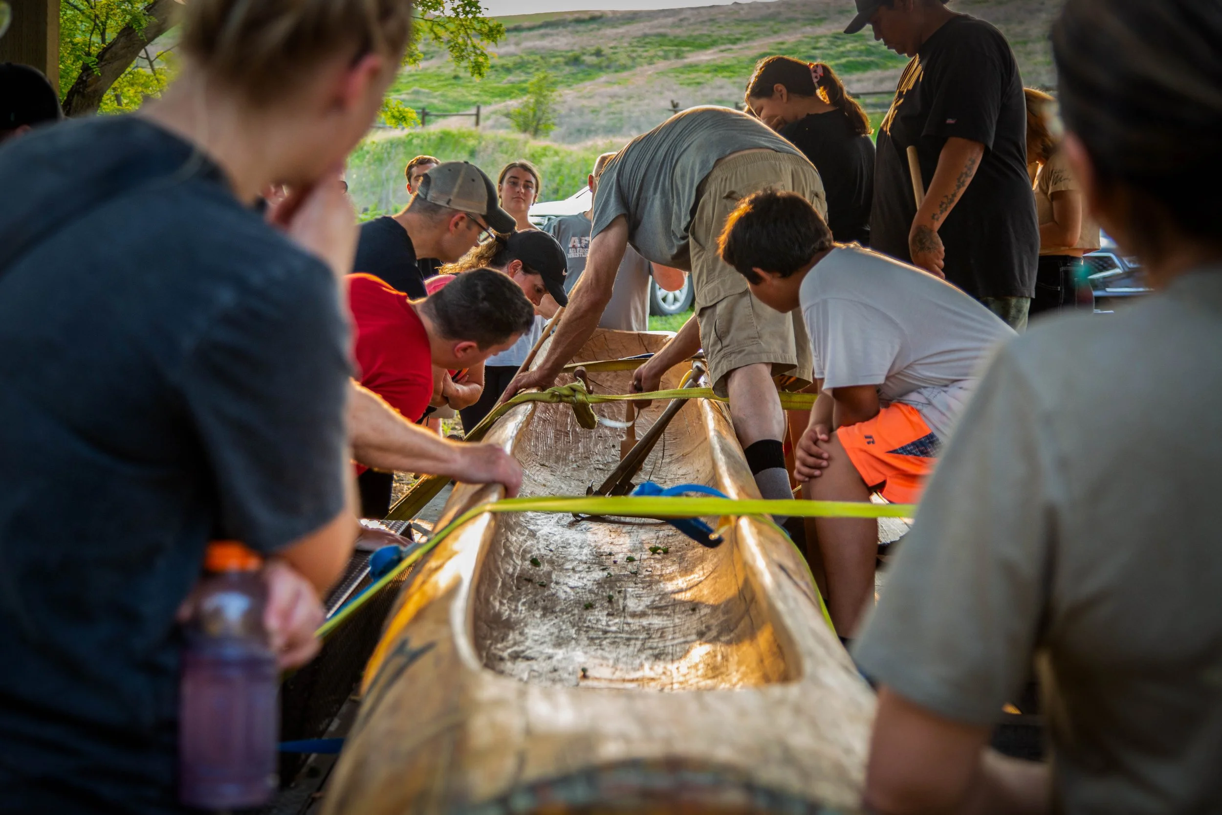 People gathered outdoors around a long wooden structure, possibly making crafts or working on a project, with a scenic green hill and trees in the background during daylight.