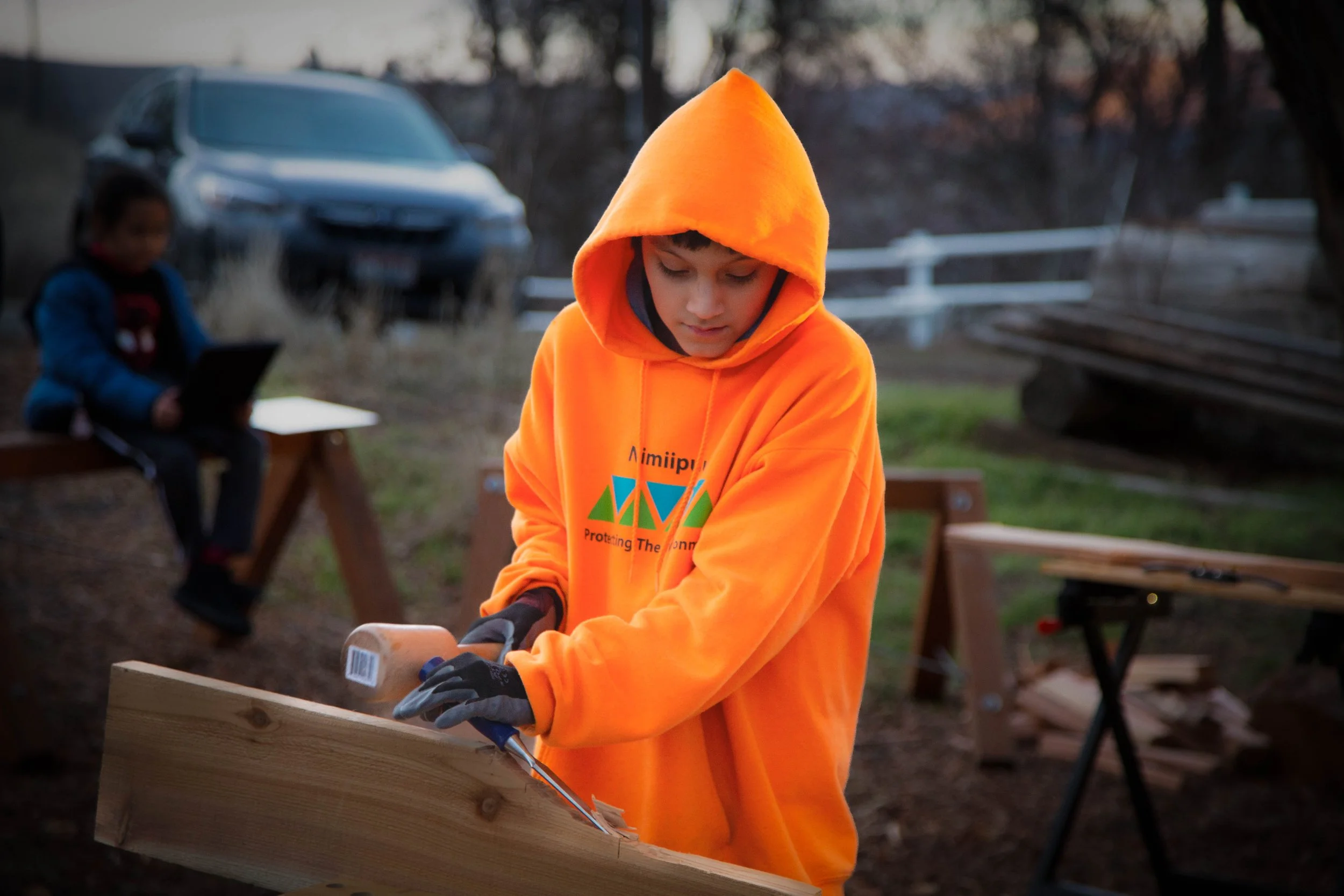 A young boy in an orange hoodie cutting wood with a handsaw outdoors during daytime, with another child sitting on a bench in the background.