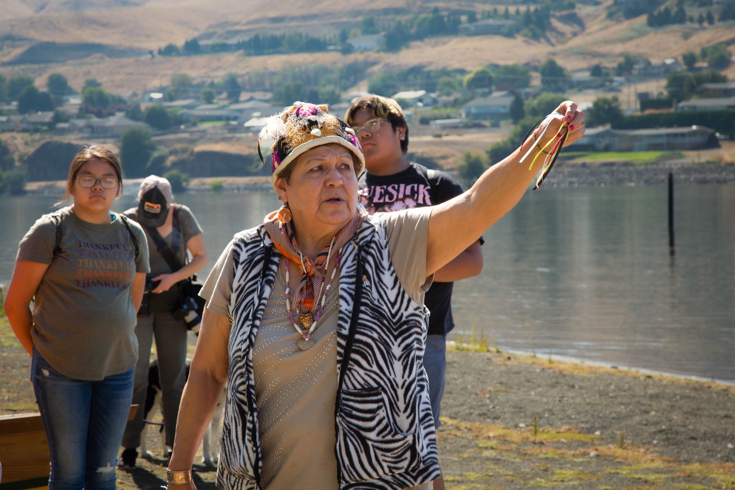 Group of people walking along a lakeside with hills and houses in the background, led by an older woman wearing a patterned vest and a colorful hat.