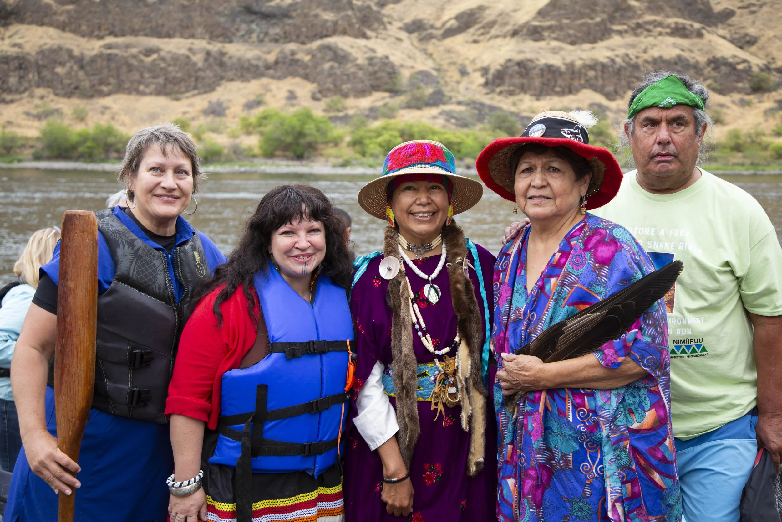 Group of five diverse people outdoors near a river, mountains in the background, dressed in colorful clothing and accessories.