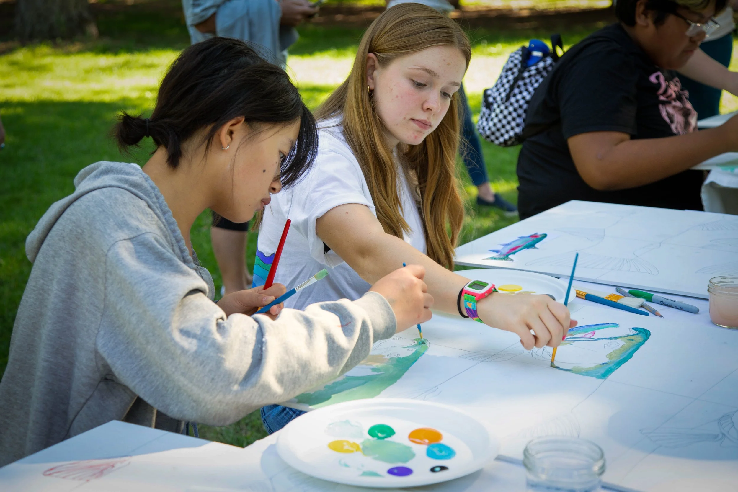 Group of children painting outdoors at a table, with paint palettes and brushes, during a sunny day.