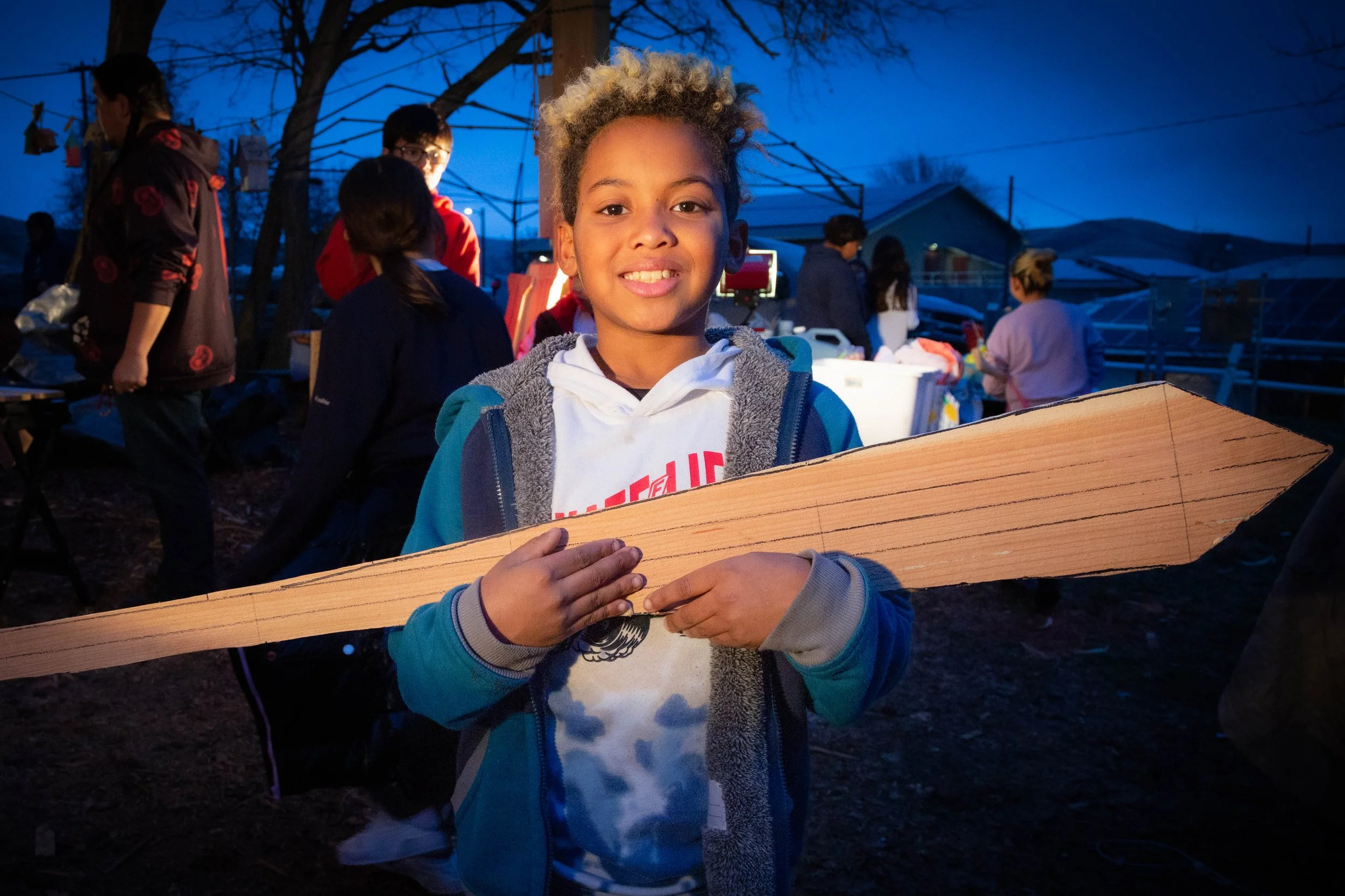 A young boy with curly blonde hair smiling at the camera while holding a wooden board, standing outdoors during dusk with other people in the background.