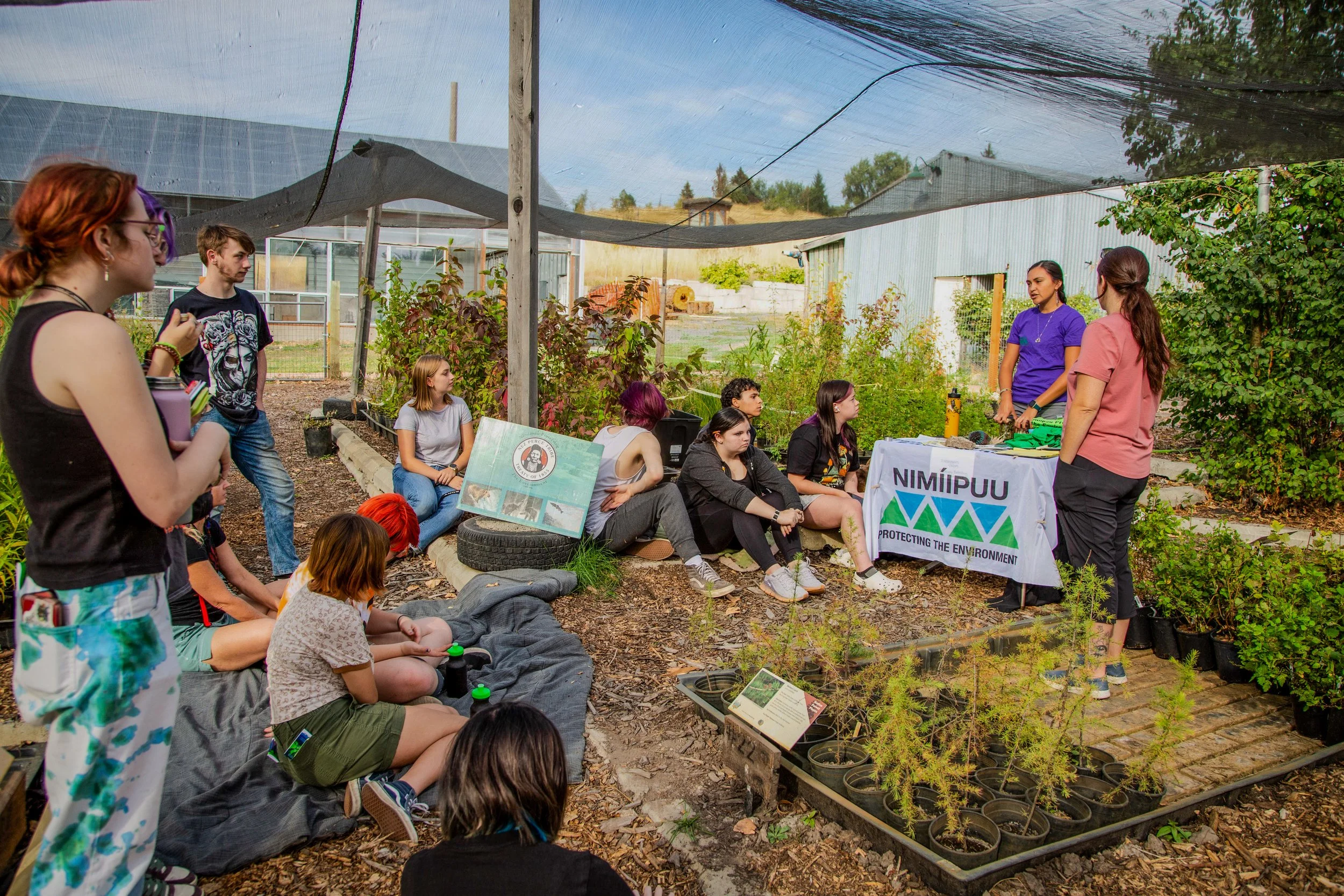 Group of young people attending an outdoor environmental education session in a garden, with plants and informational displays, one person speaking at a table with a banner that says 'Protecting the Environment' in front of lush greenery.