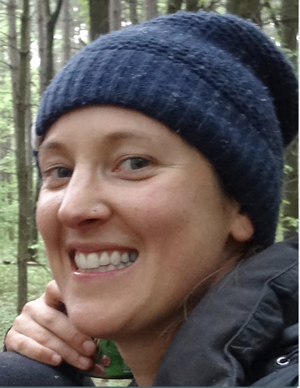 Close-up of a woman smiling outdoors in a forest, wearing a dark blue knit beanie and black jacket.