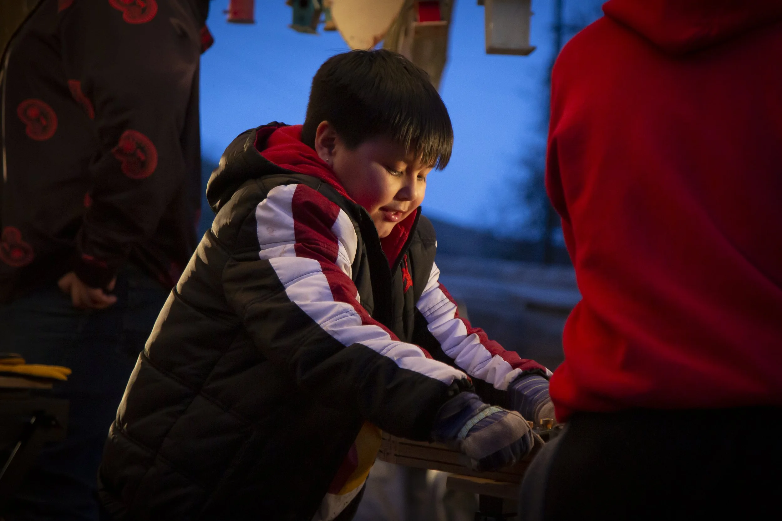A young boy wearing a winter jacket and gloves, focusing on a task in an outdoor setting during evening or dusk, with other people partially visible around him.